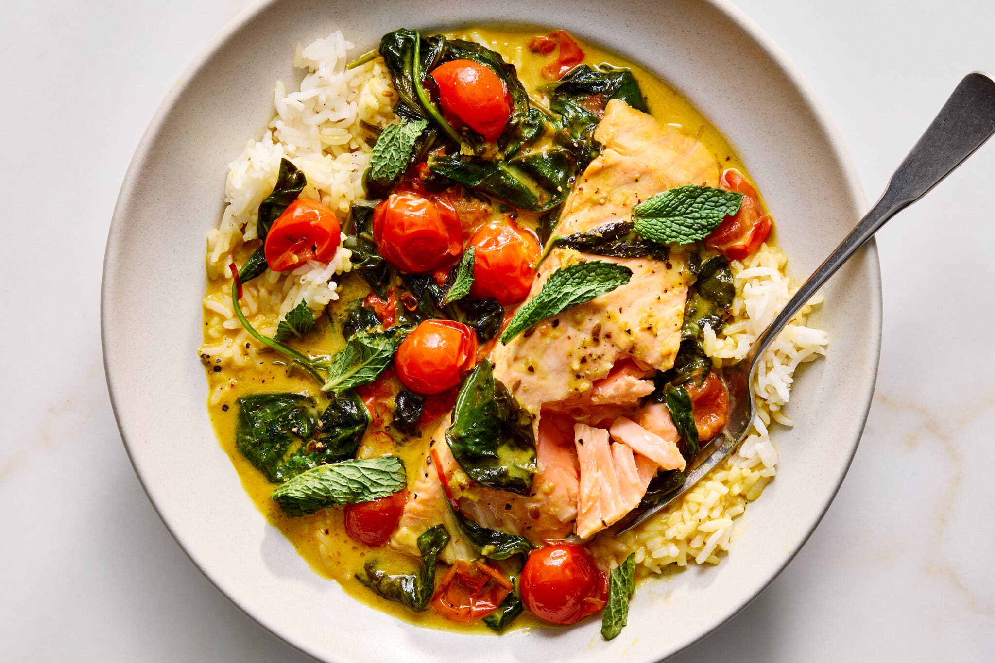 A salmon fillet served in a yellow coconut curry broth with wilted spinach, cherry tomatoes and white rice.