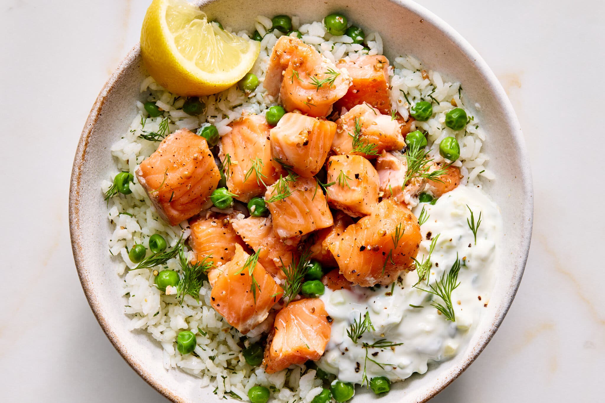 Chunky pieces of pink salmon over white rice with green peas, fresh dill, white sauce and a lemon wedge.