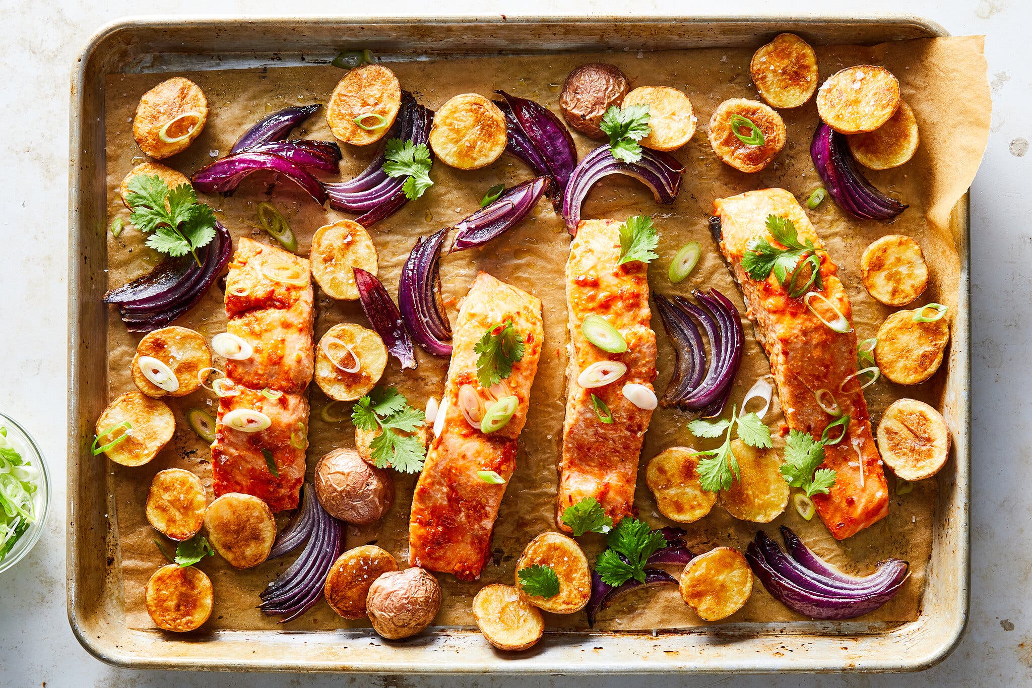 Four glazed salmon fillets roasted on a sheet pan with sliced potatoes, red onion wedges and fresh cilantro.