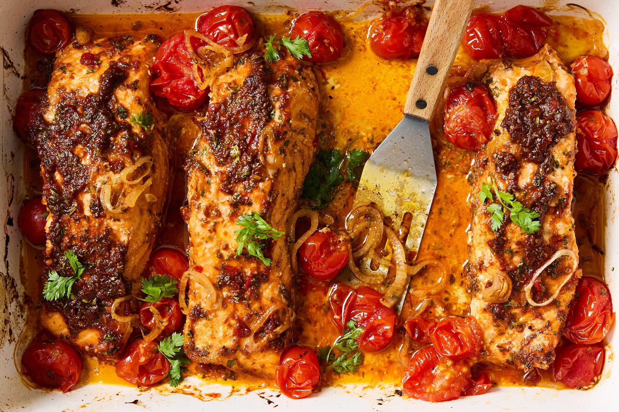 Three salmon fillets roasted in a baking dish with dark red harissa paste, blistered cherry tomatoes and sliced shallots. 