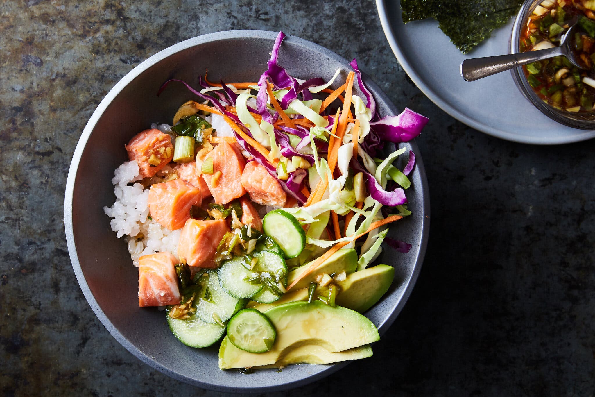 A gray bowl filled with cubed salmon over white rice, topped with sliced cucumbers, avocado and shredded carrots and cabbage.