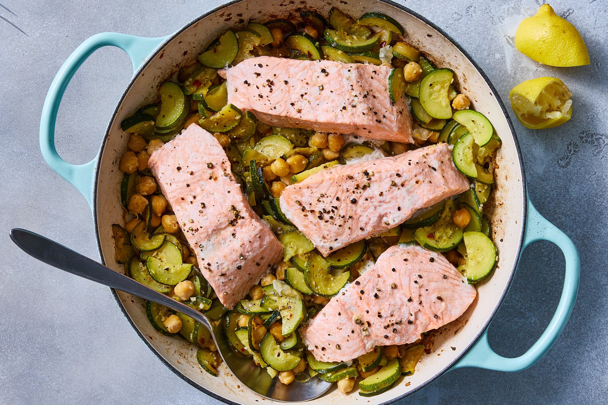 Four salmon fillets cooked in a light blue pot over a bed of sliced zucchini and chickpeas.