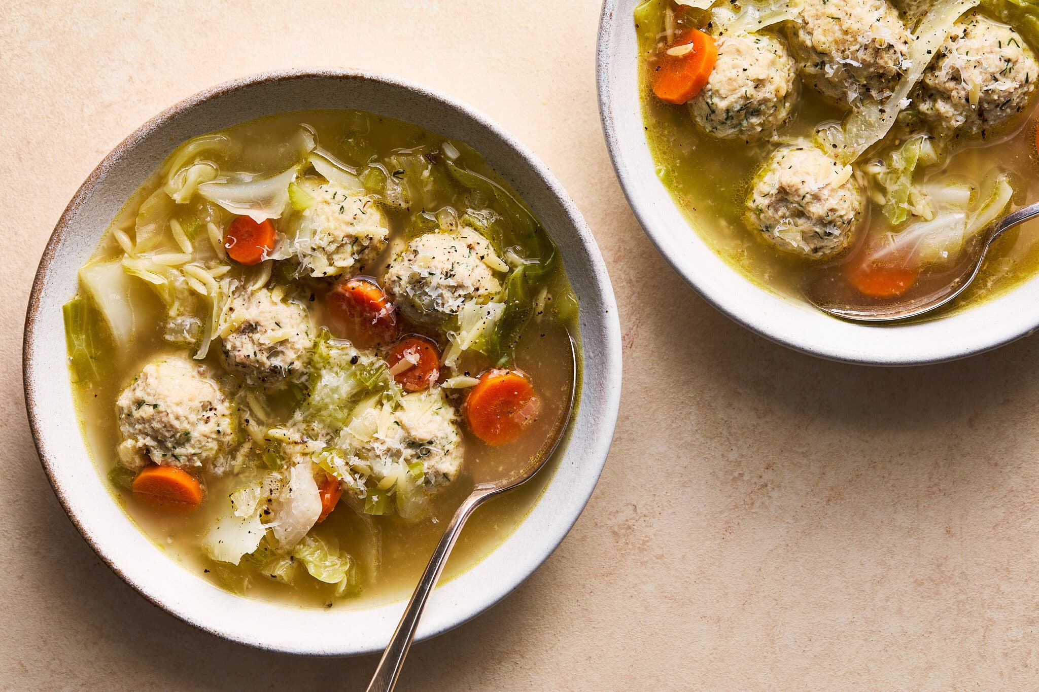 An overhead image of two bowls of chicken soup: meatballs and cabbage float in a golden broth.