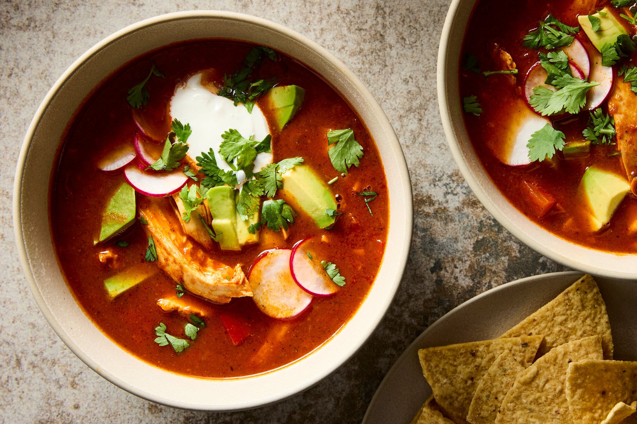 An overhead image of a chicken in a bright red broth, finished with cilantro, avocado, radishes and a dollop of sour cream.