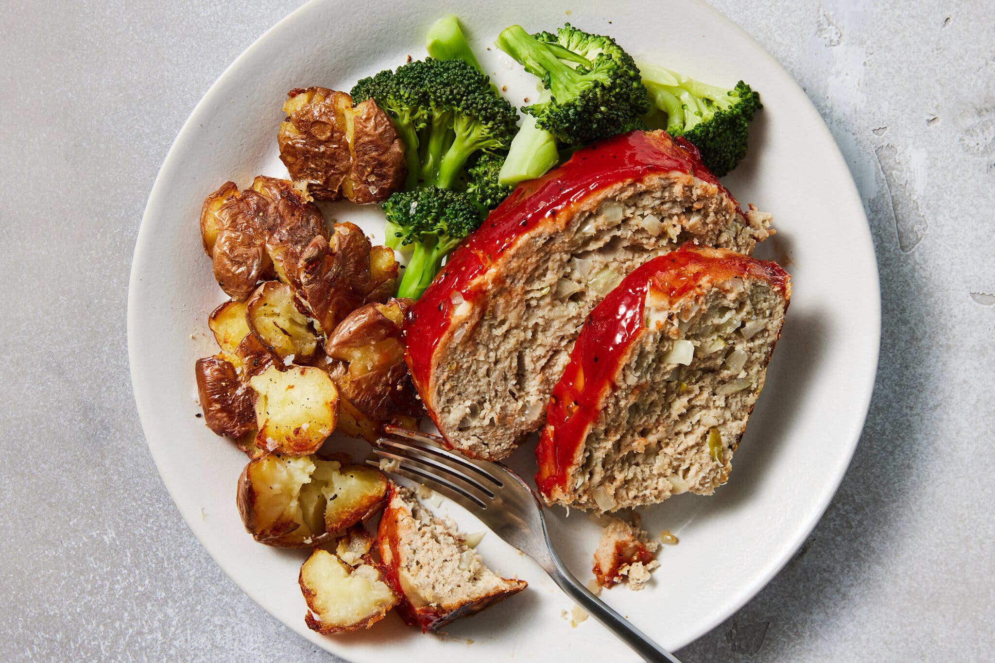 An overhead image of a plate of meatloaf with a ketchup glaze, broccoli and smashed roasted potatoes.
