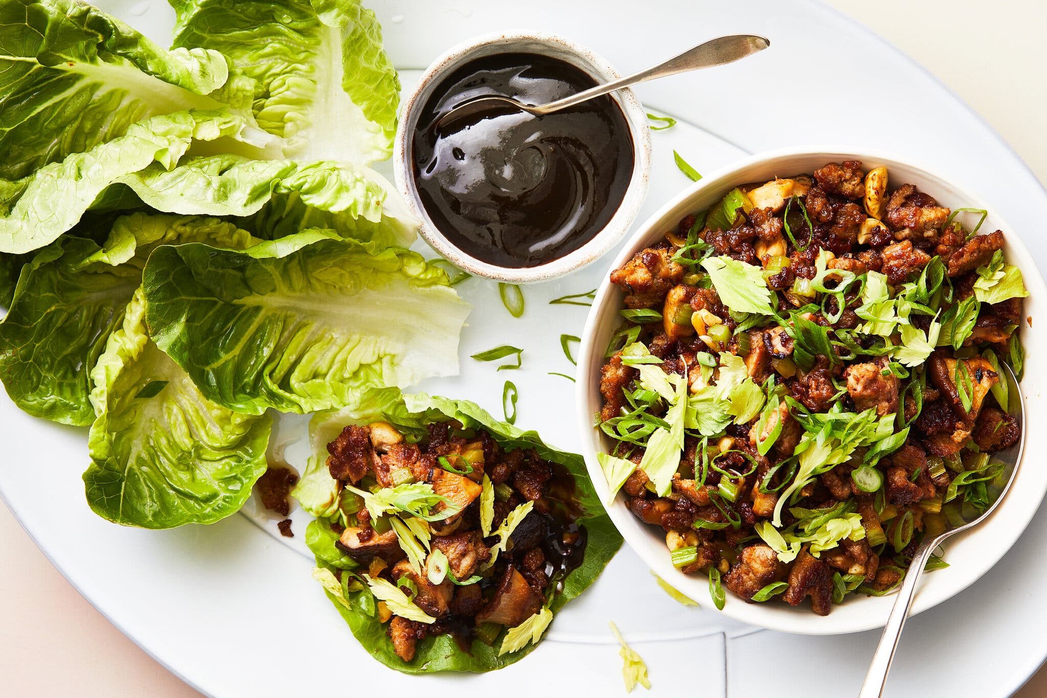 An overhead image of a tray of food including lettuce leaves, a bowl of hoisin sauce with a spoon in it and a bowl of diced cooked chicken and vegetables garnished with cilantro and scallions. One lettuce cup is constructed and sitting beside the ingredients.