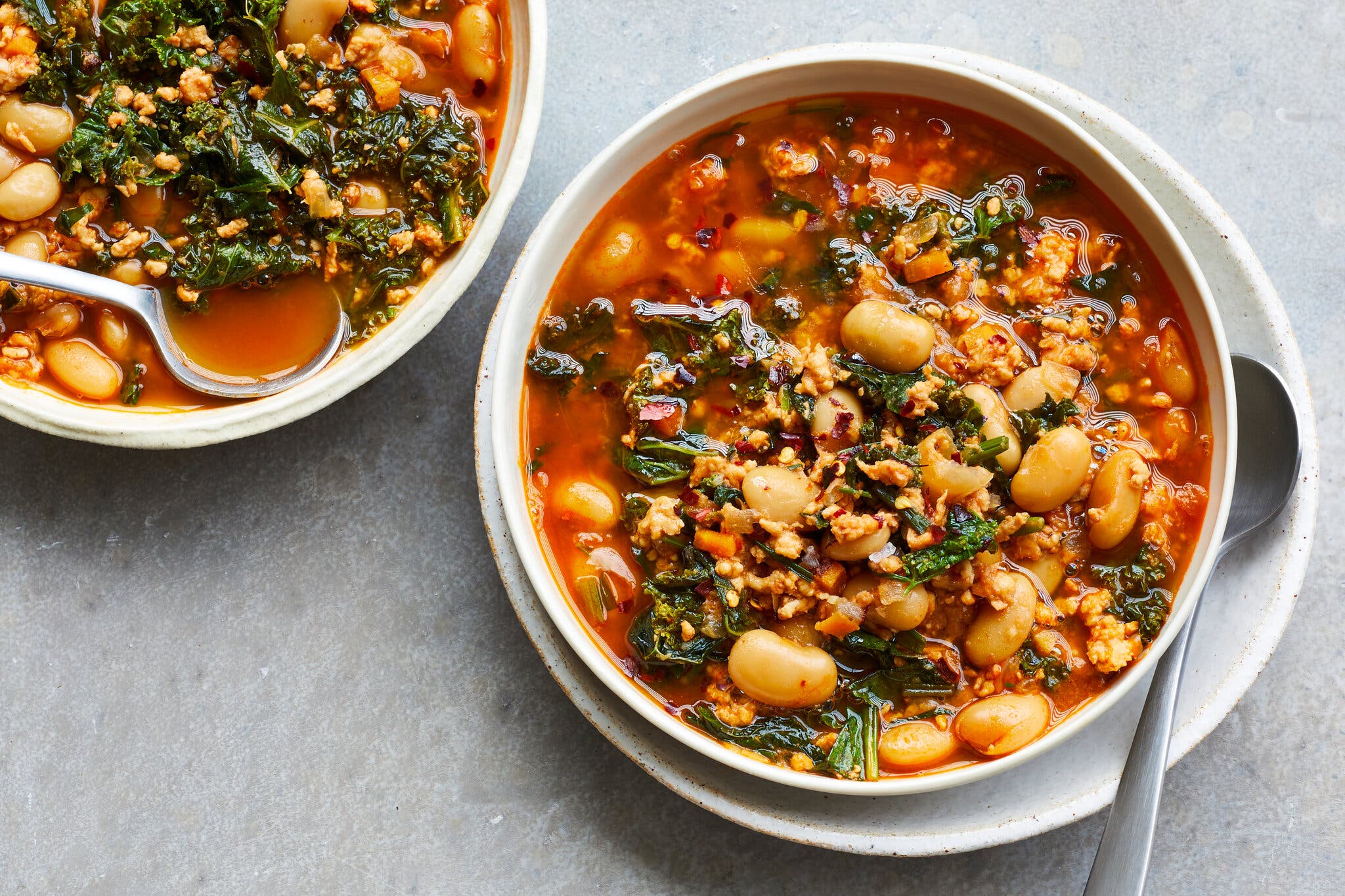 An overhead image of a bowl of tomatoey soup with ground turkey, white beans and kale on a plate, with a spoon. There is another bowl of soup in the top left corner.