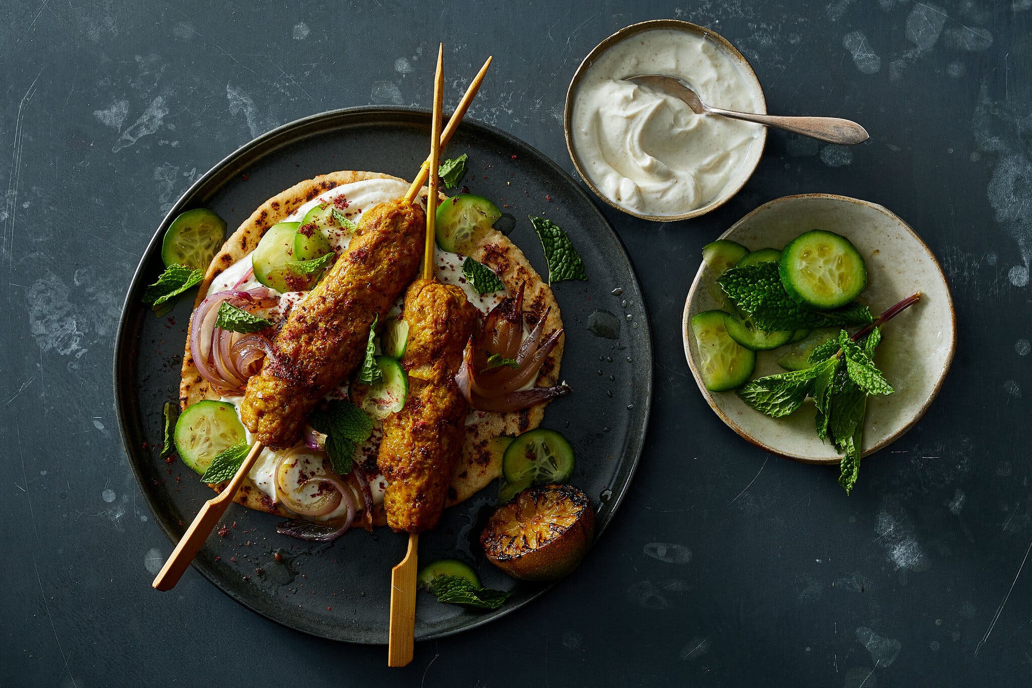 On a dark marble counter, a plate of two wooden skewers of turkey kofta, lying on top of two pitas smeared with yogurt sauce and cucumbers, garnished with mint. There is a bowl of yogurt sauce and a small plate of cucumbers and mint nearby.