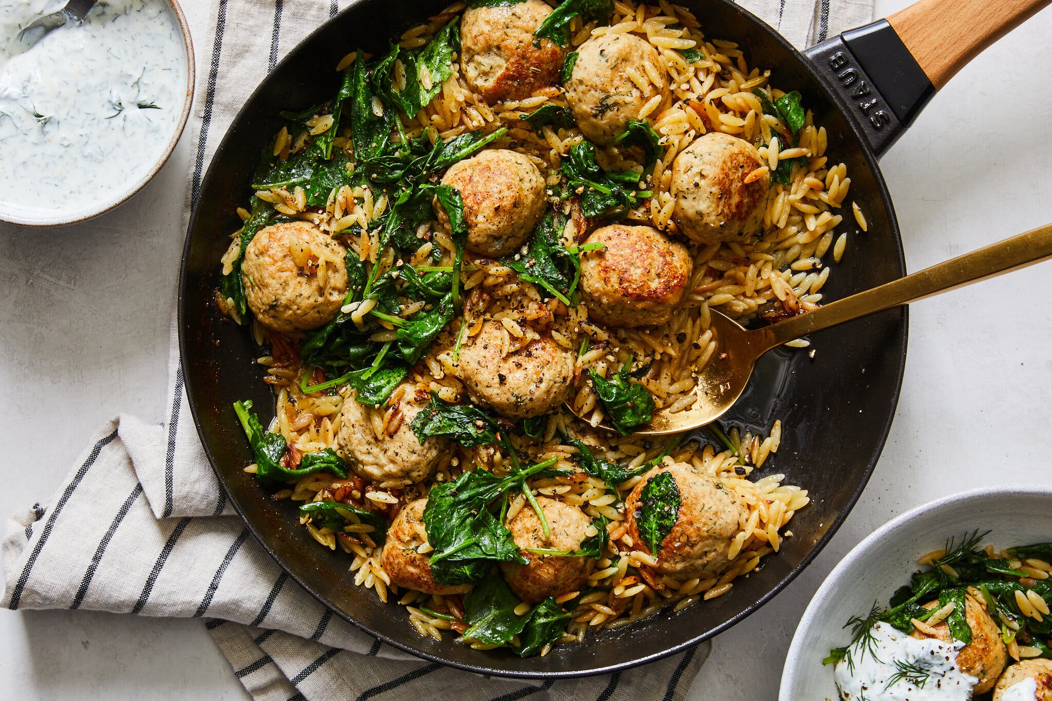 An overhead image of a skillet full of orzo with greens and turkey meatballs, set on top of a dish towel. There is a spoon in the skillet.