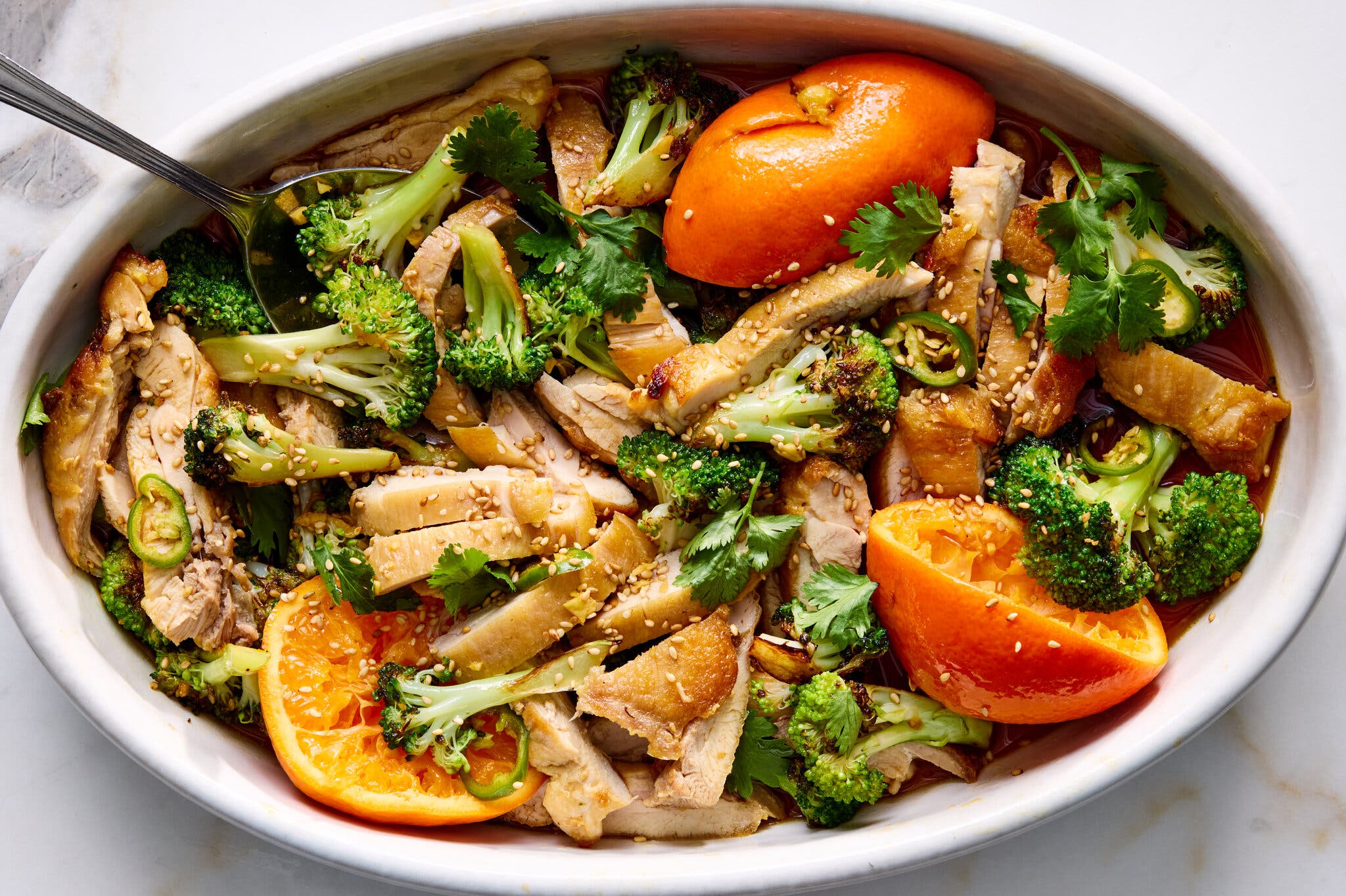 Sliced roasted chicken and broccoli florets in a white dish garnished with orange halves, cilantro and sesame seeds.