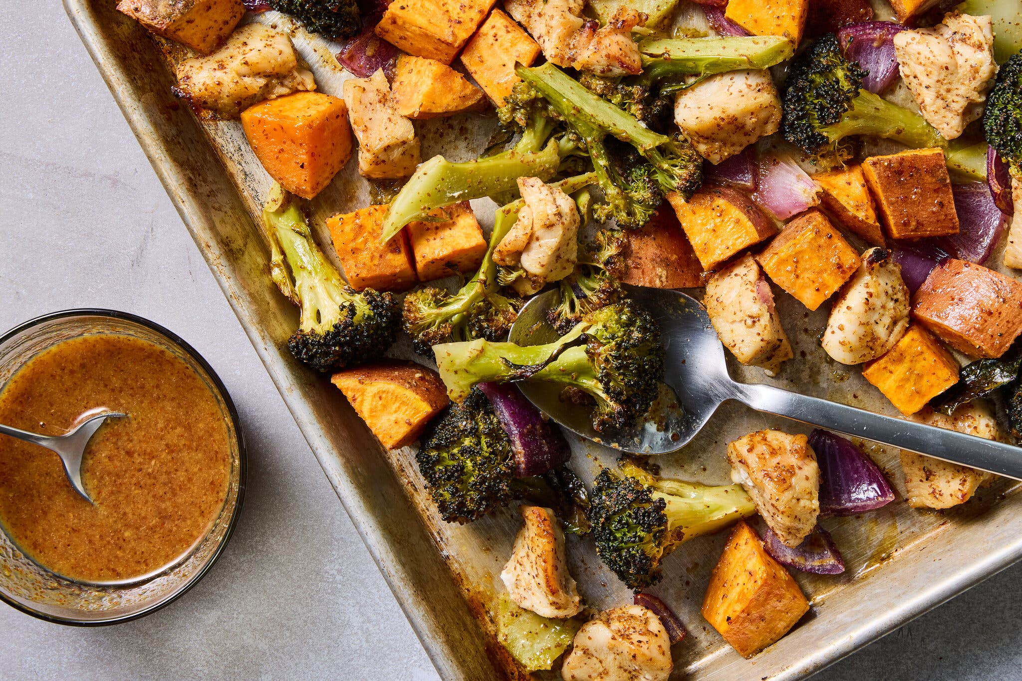 Roasted chicken, sweet potatoes, broccoli and red onion on a baking sheet next to a bowl of hot honey.