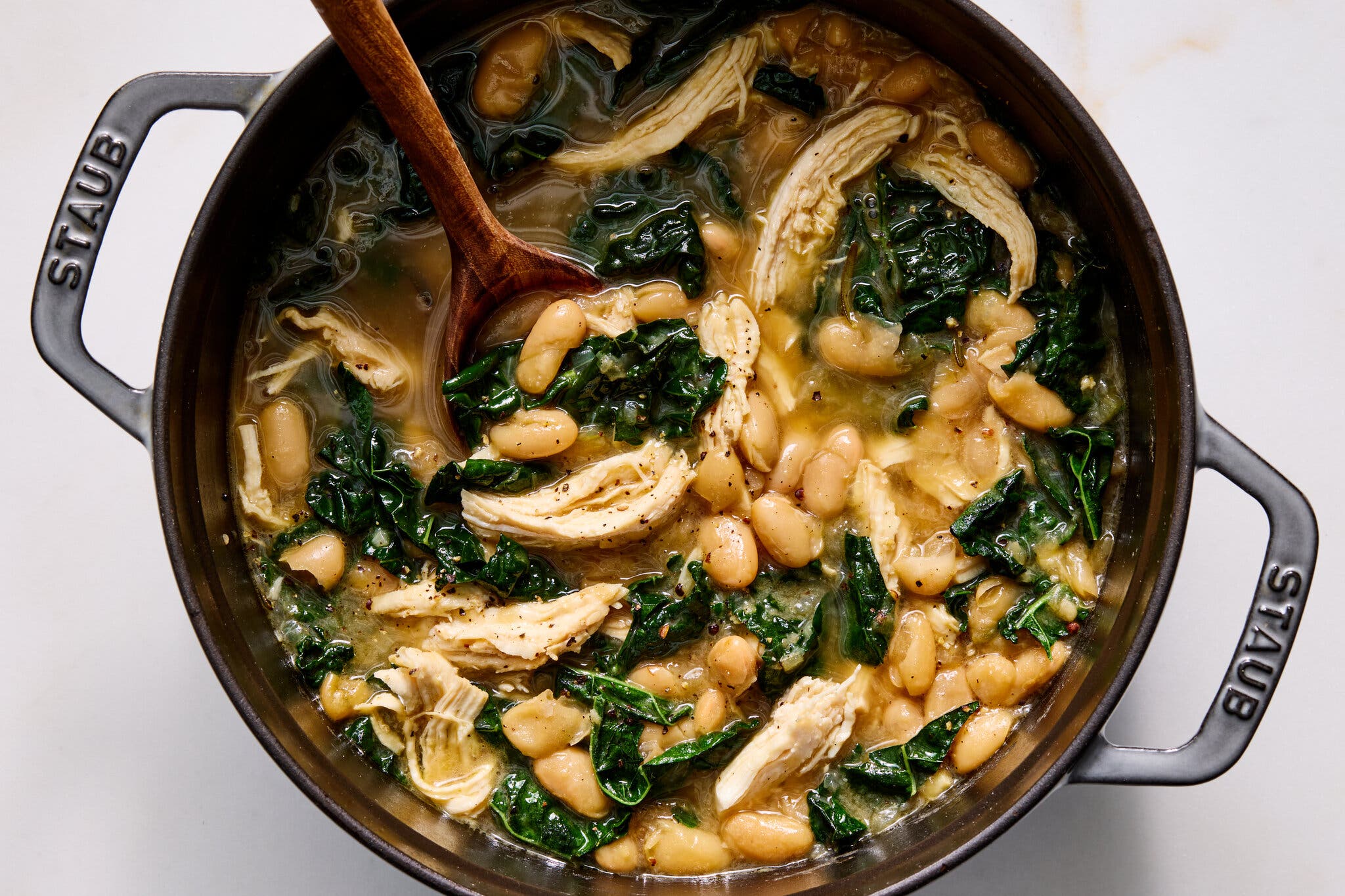 Shredded chicken, white beans and dark leafy kale in a savory broth inside a black Staub pot.