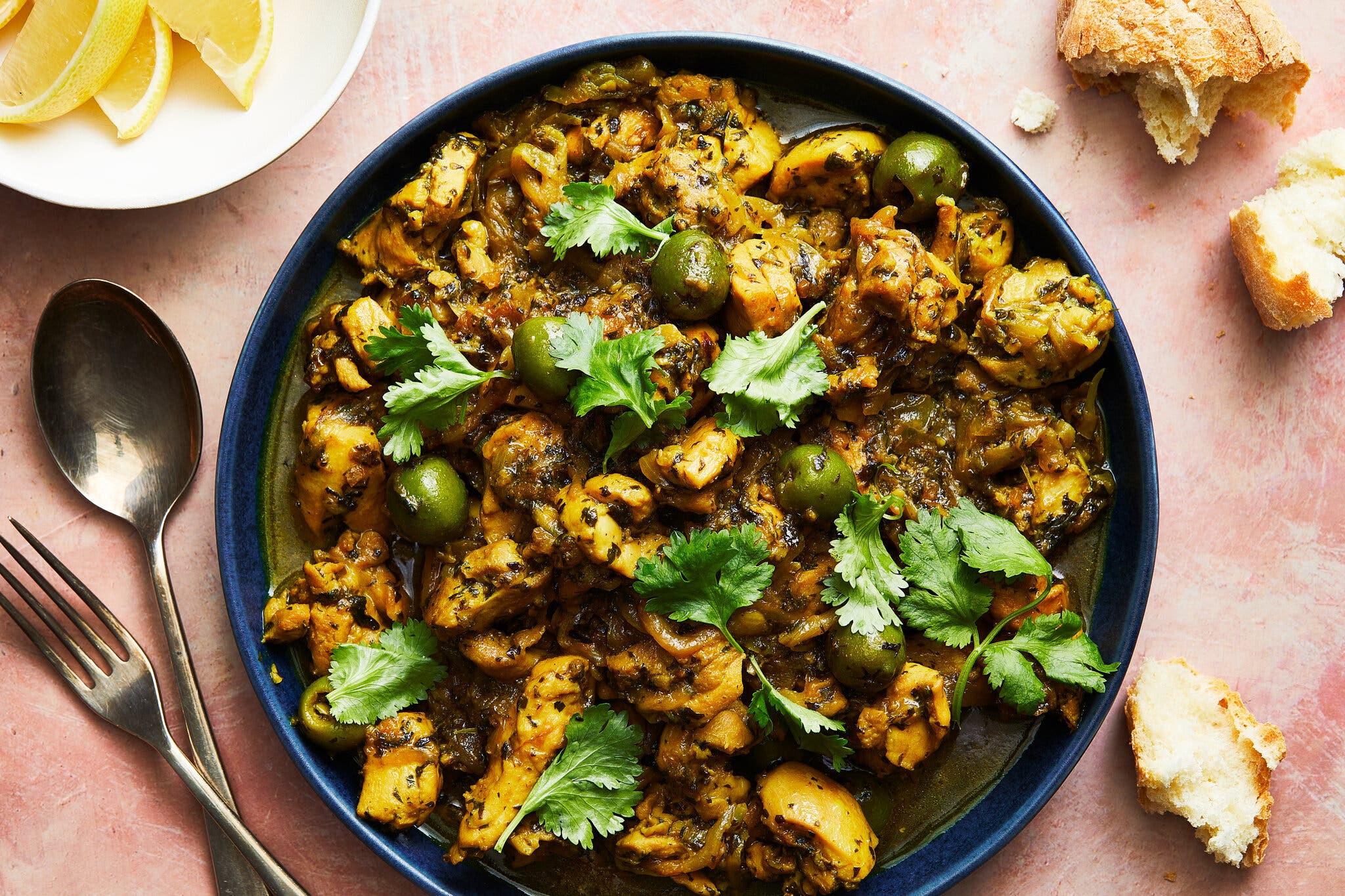 A blue bowl of chicken stew with green olives and fresh cilantro, served with crusty bread.