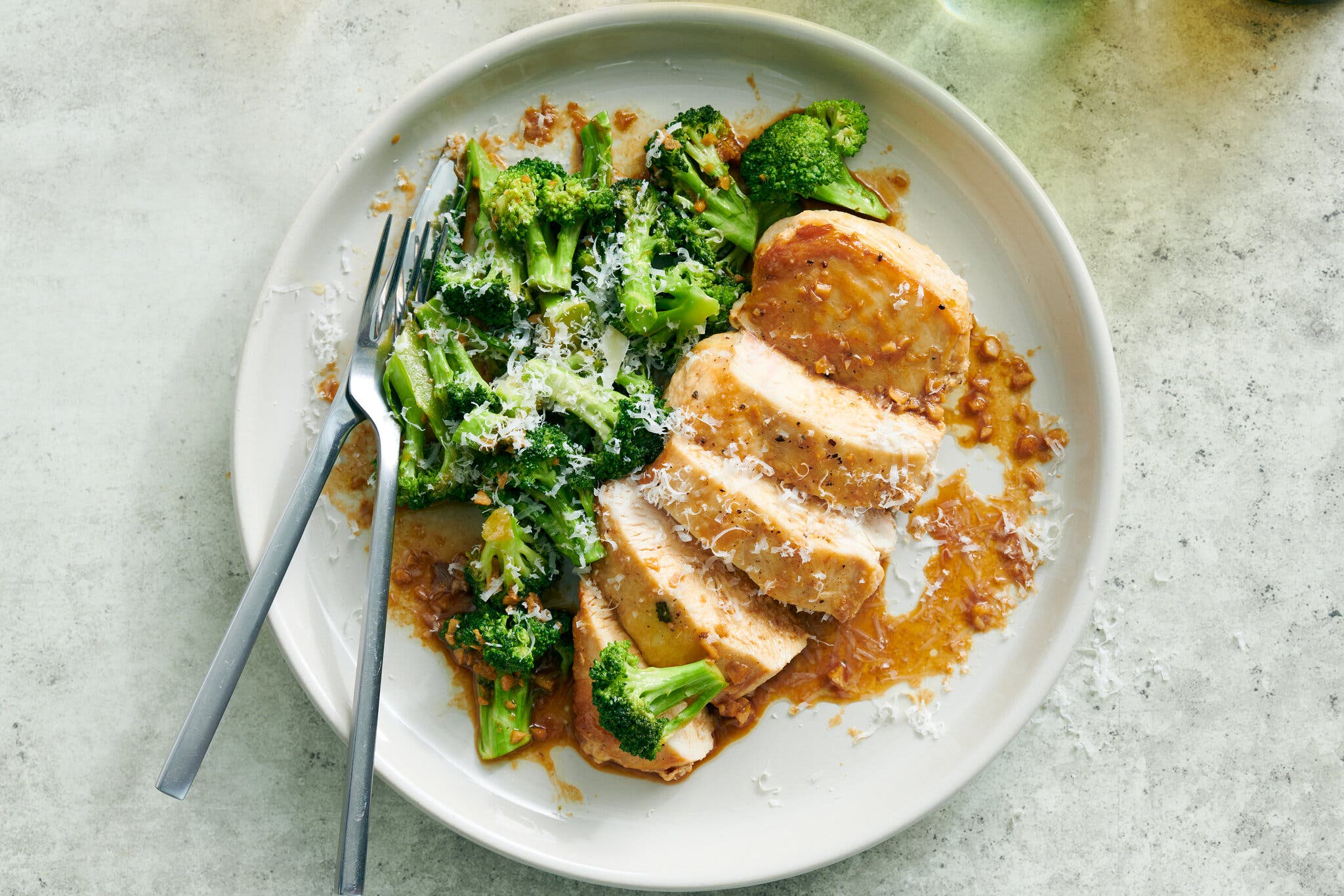 Sliced chicken breast and steamed broccoli topped with a golden sauce and grated Parmesan on a white plate.