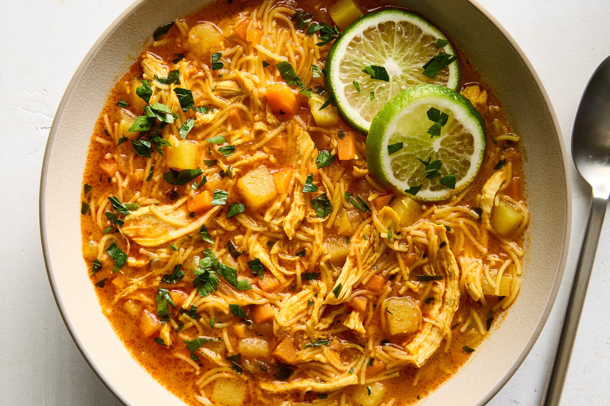 A bowl of golden-orange soup with shredded chicken, diced potatoes and vermicelli noodles and garnished with lime wheels.