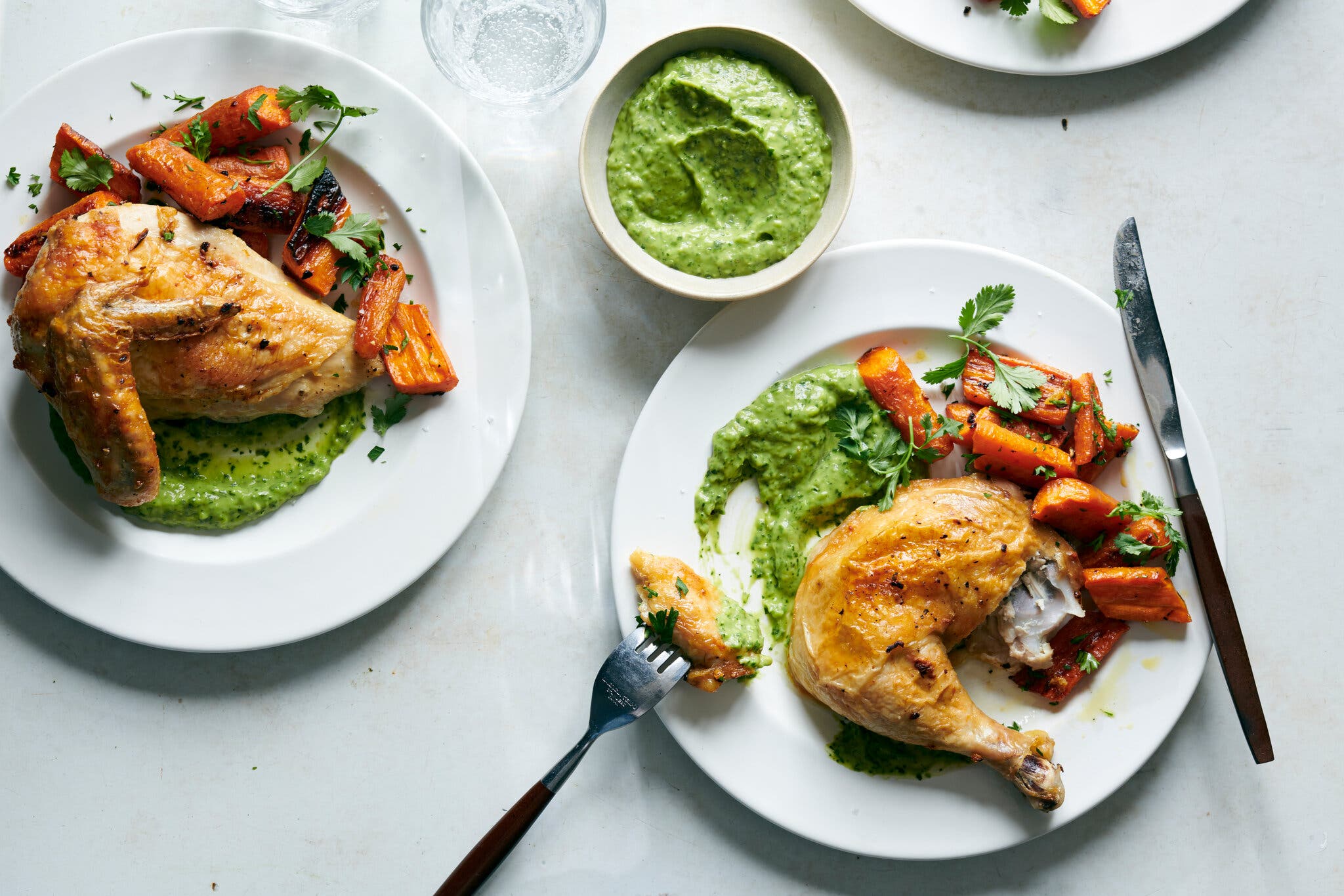 Two plates of roasted chicken and carrots served with a vibrant green herb sauce.