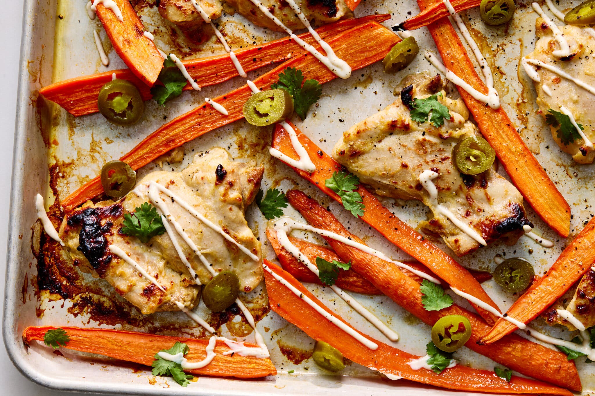 Roasted chicken thighs, long carrot halves and pickled jalapeño coins drizzled with a creamy white sauce on a sheet pan.