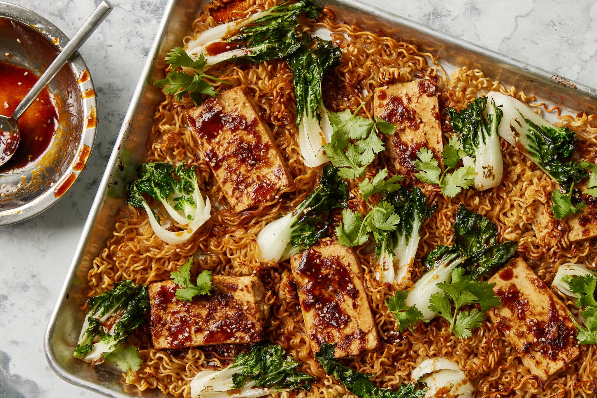 Crispy glazed tofu blocks and bok choy arranged over a bed of ramen noodles on a sheet pan.