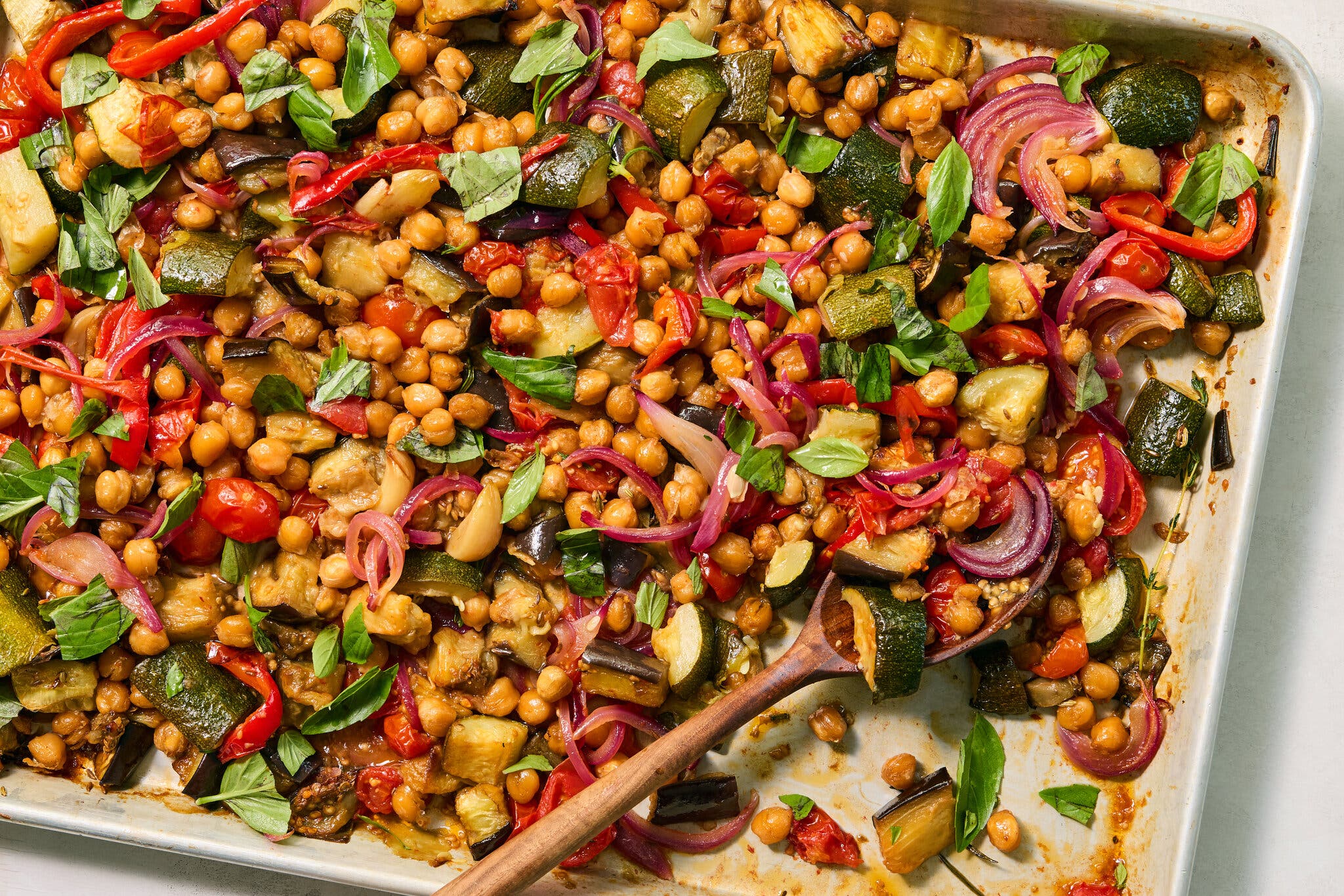 A crowded sheet pan of roasted chickpeas, zucchini, red onions and tomatoes topped with fresh basil leaves.