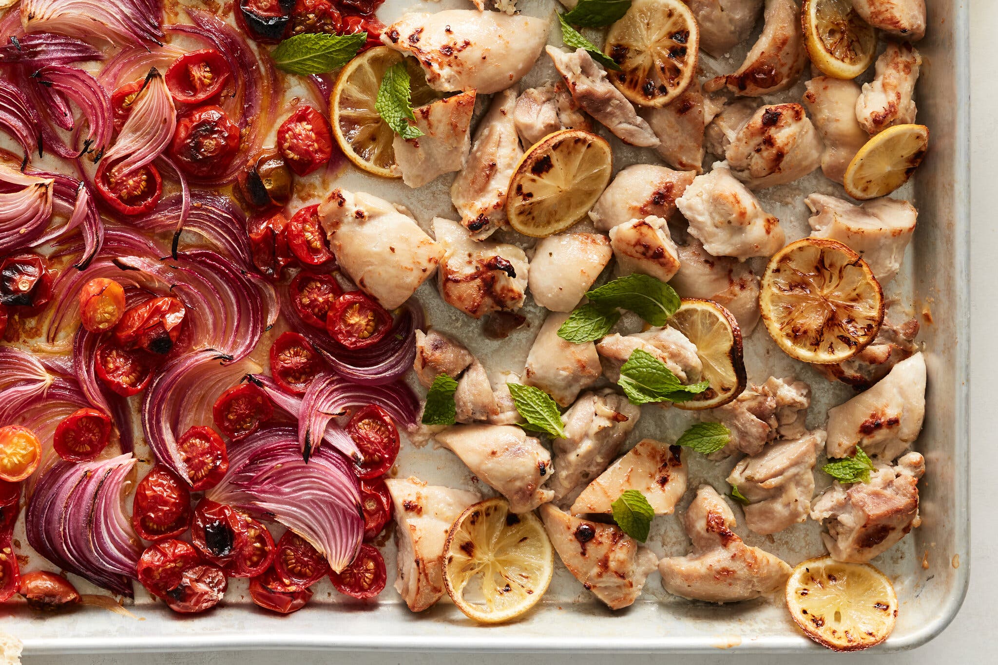 Roasted boneless chicken pieces, charred lemon wheels, red onion wedges and cherry tomatoes on a sheet pan.