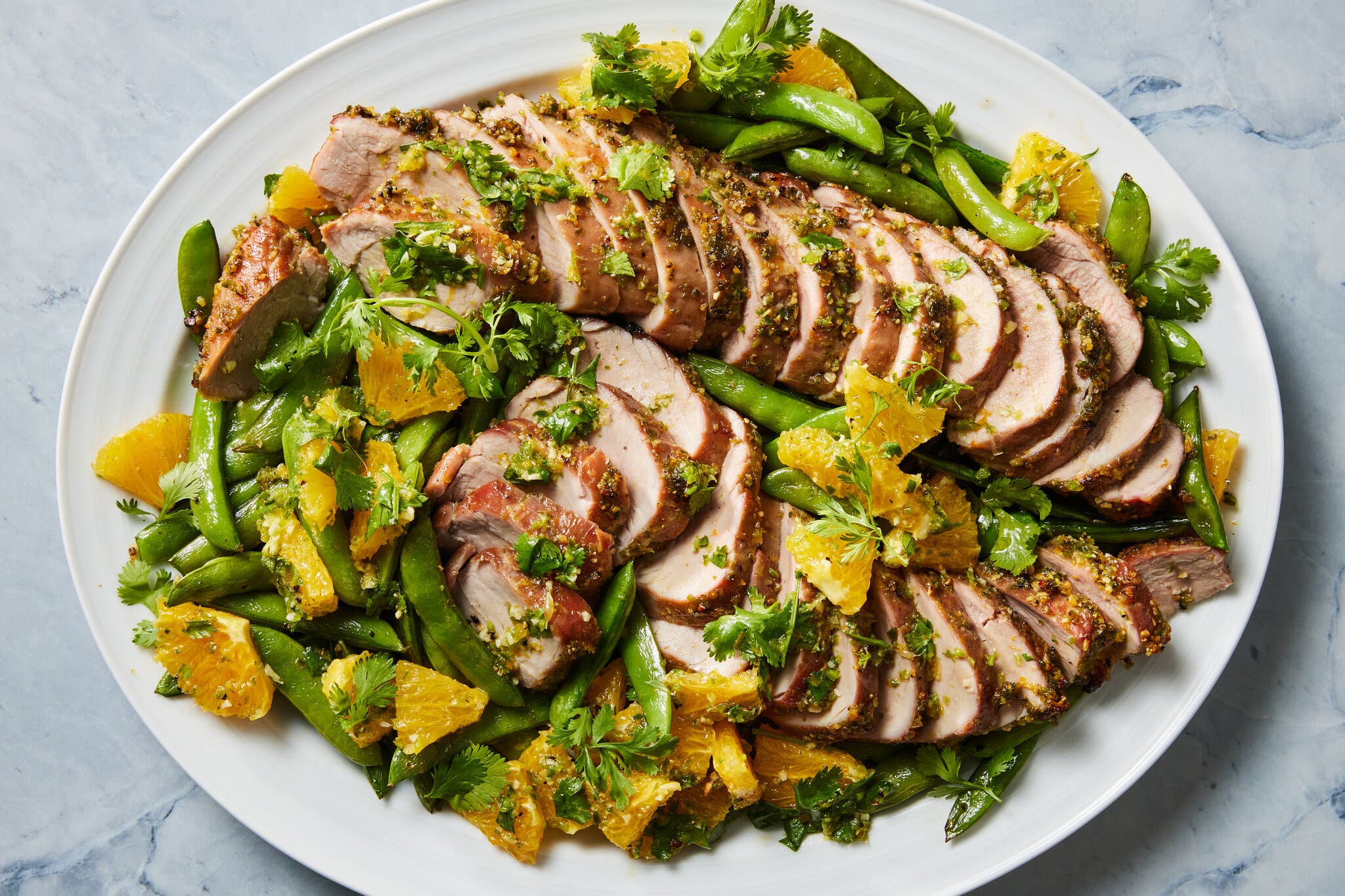 Sliced pork tenderloin arranged on a platter with sugar snap peas, orange segments and a herb sauce.