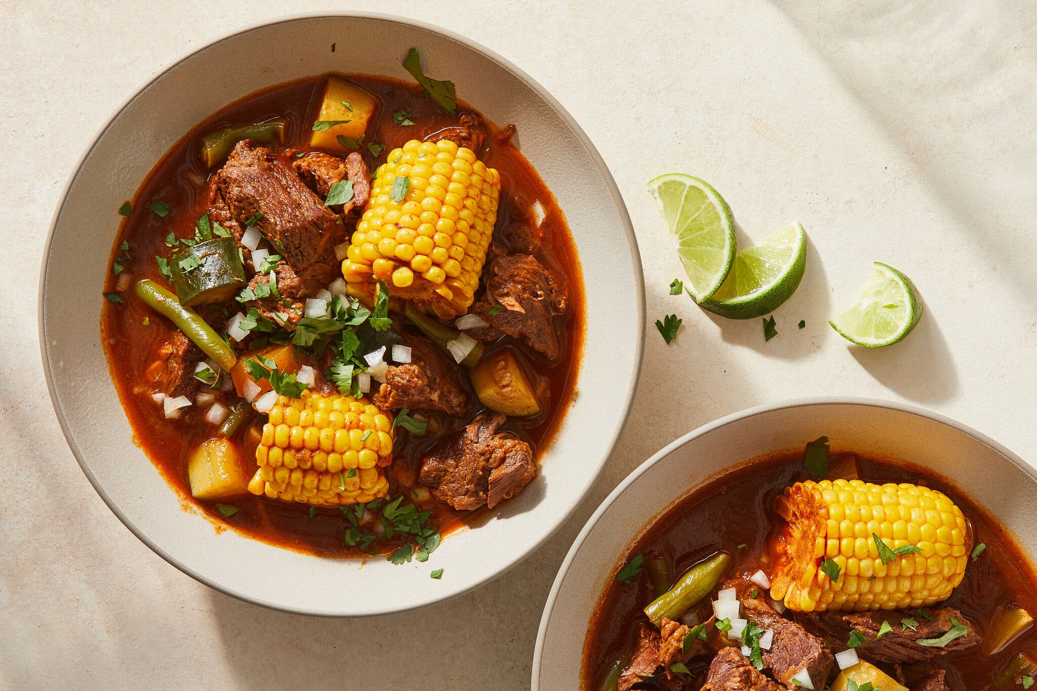 Two bowls of reddish-brown beef stew featuring large sections of yellow corn on the cob and lime wedges.