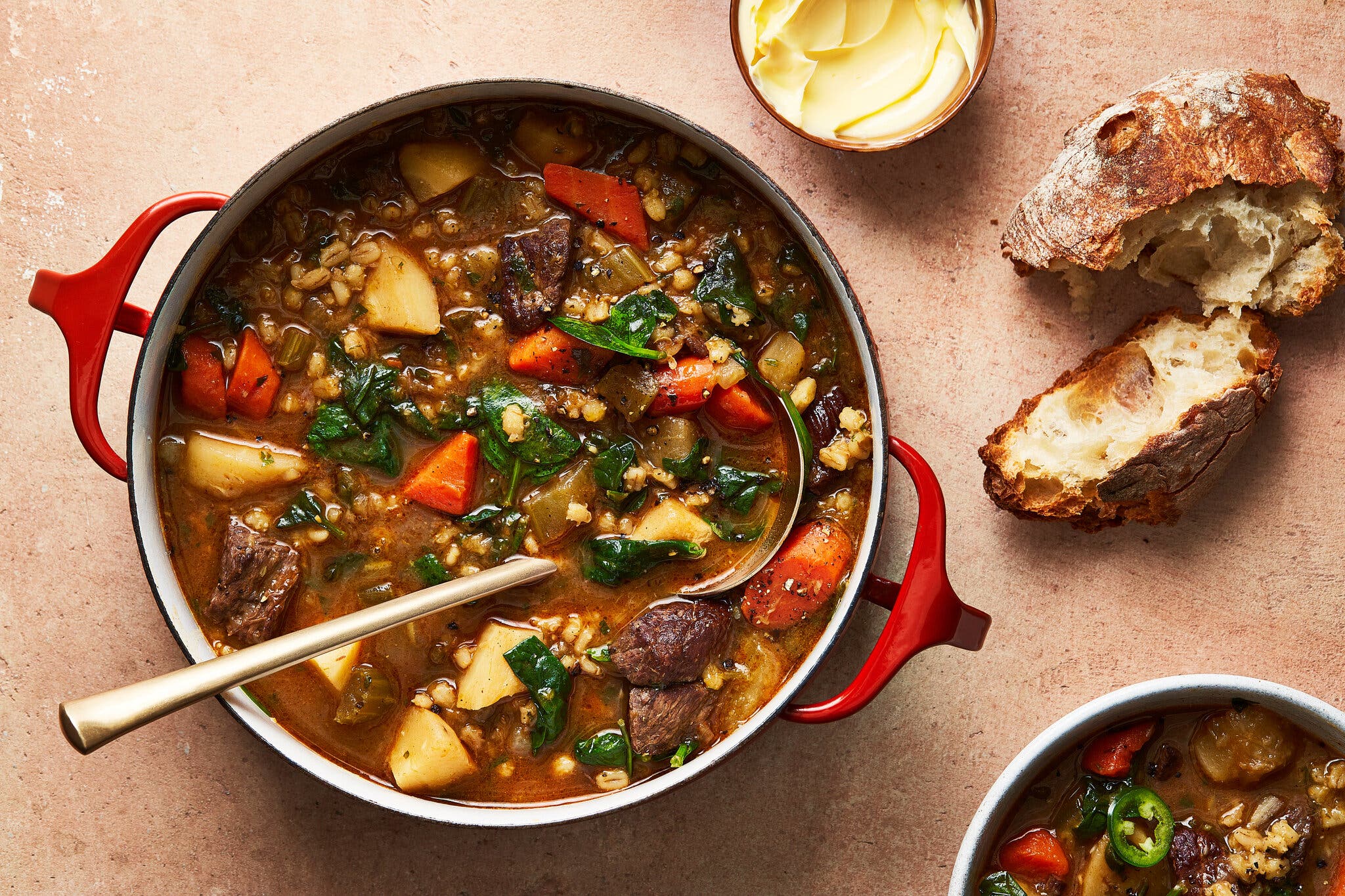 A red Dutch oven filled with beef barley stew and spinach, served with crusty bread and butter.