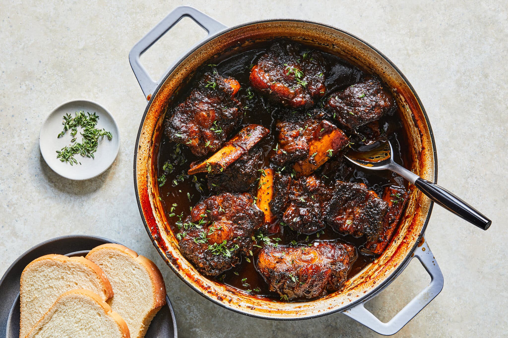 Bone-in short ribs simmer in a rich, nearly black sauce inside a blue pot. A small plate of thyme and slices of white bread are arranged on the side.