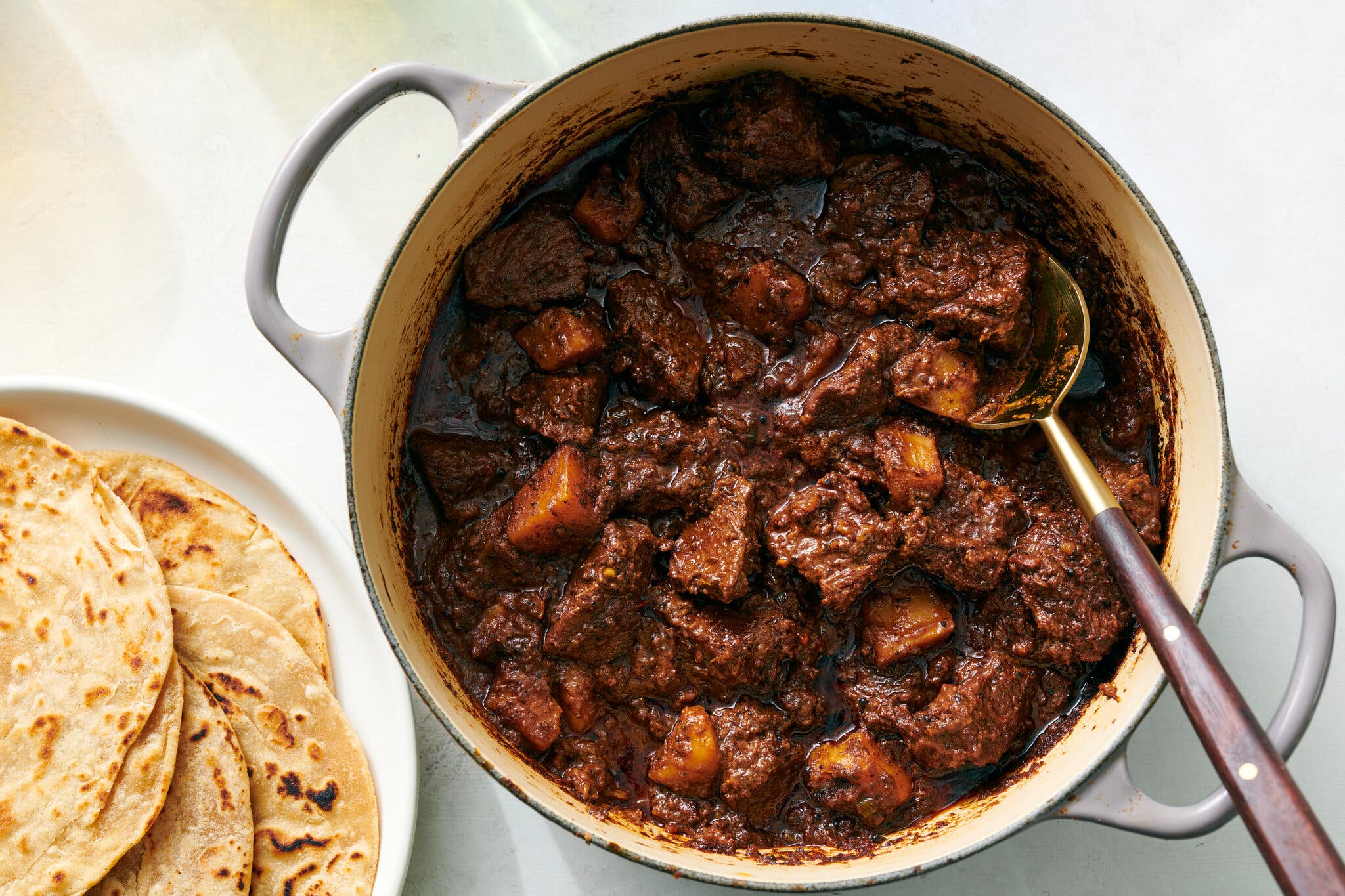 Chunks of beef and potatoes in a thick, dark brown sauce sit in a light gray pot with a gold and wooden serving spoon. A stack of charred flatbread sits on a white plate.