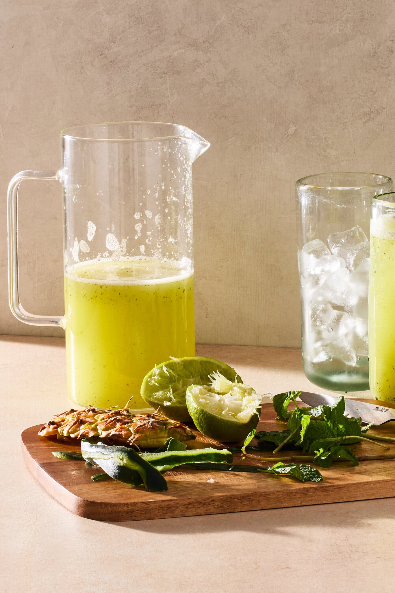 A pitcher of a yellow drink sits beside a cutting board with pineapple and lime rinds, cucumber peels and mint sprigs.