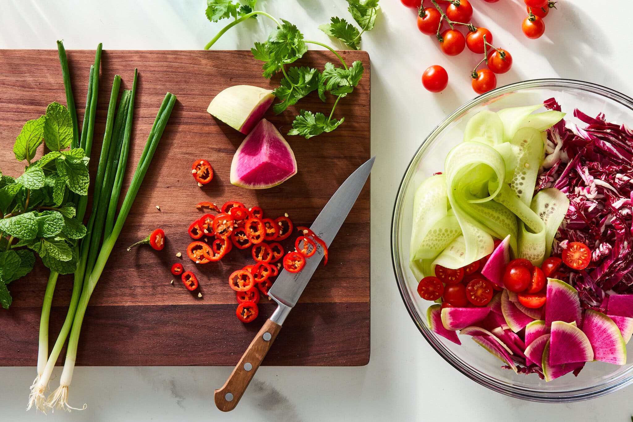 An overhead image of ingredients on a cutting board next to a glass bowl full of cut cucumbers, tomatoes, radishes and cabbage.