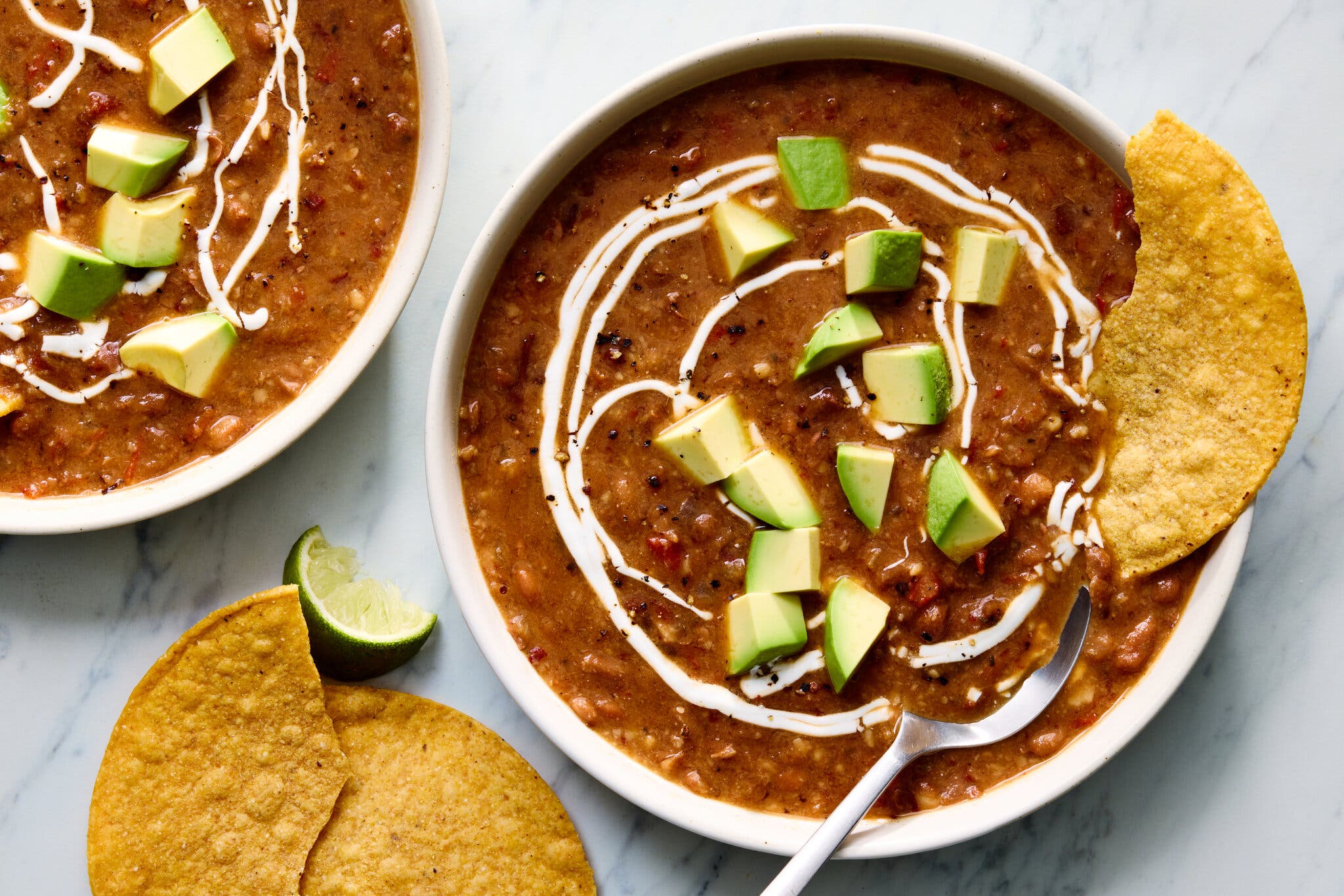 Creamy bean soup in a white bowl topped with avocado cubes, a cream drizzle and a side of tortilla chips.