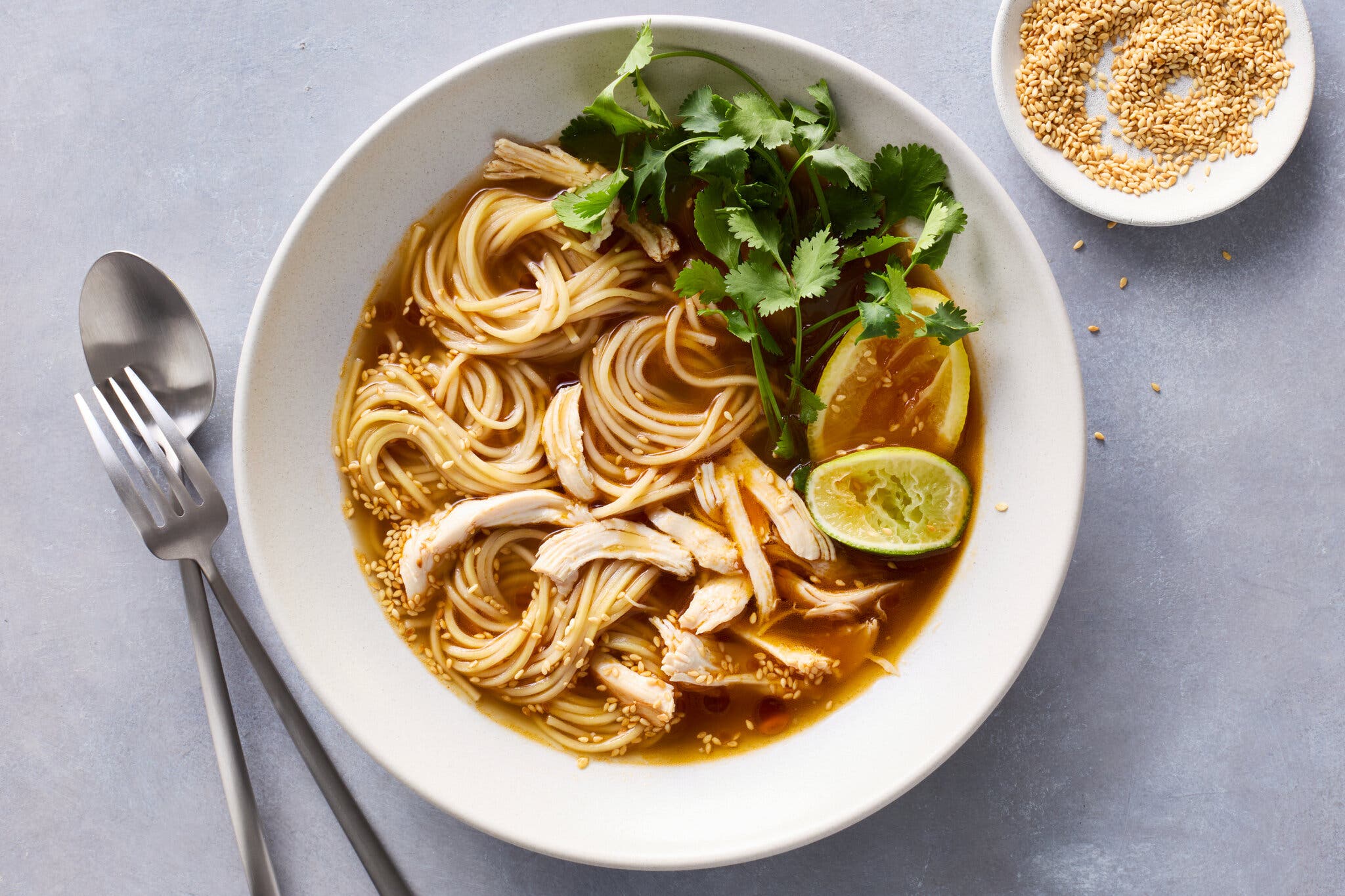 A bowl of chicken noodle soup in a dark broth topped with cilantro, lime and a sprinkle of sesame seeds.