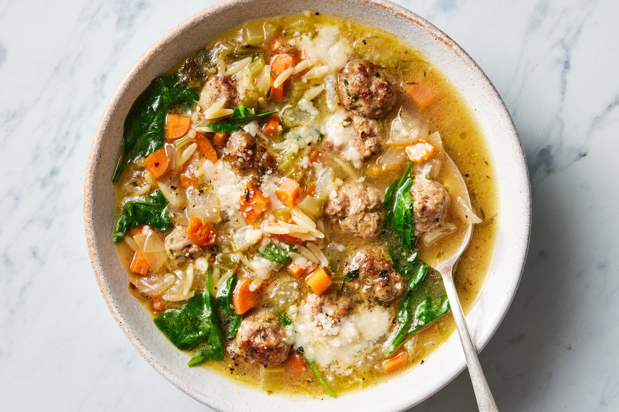 A bowl of broth with small meatballs, orzo pasta, spinach, carrots and a dusting of cheese.