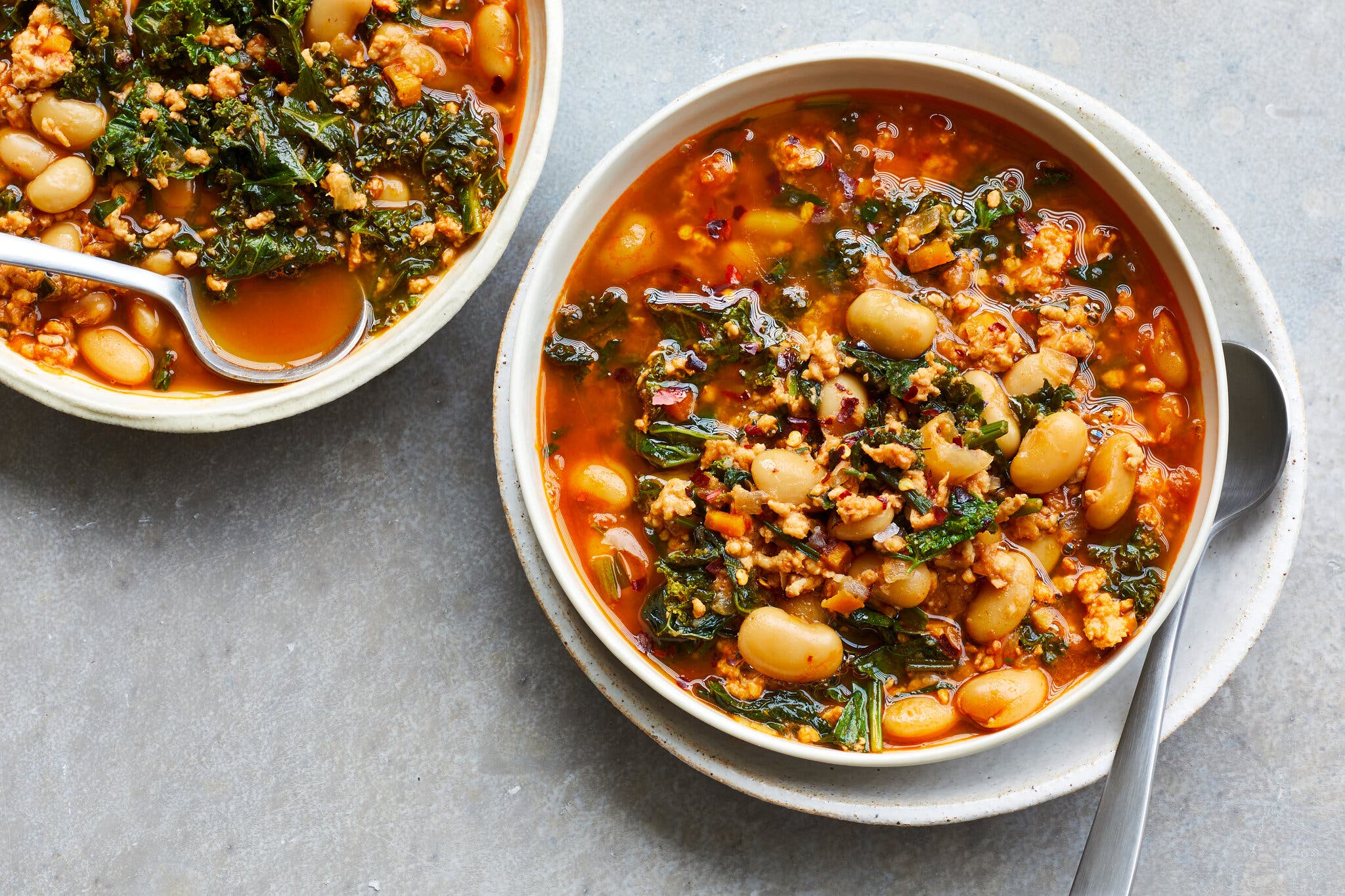 Two bowls of thick reddish stew with white beans, crumbled sausage and kale.