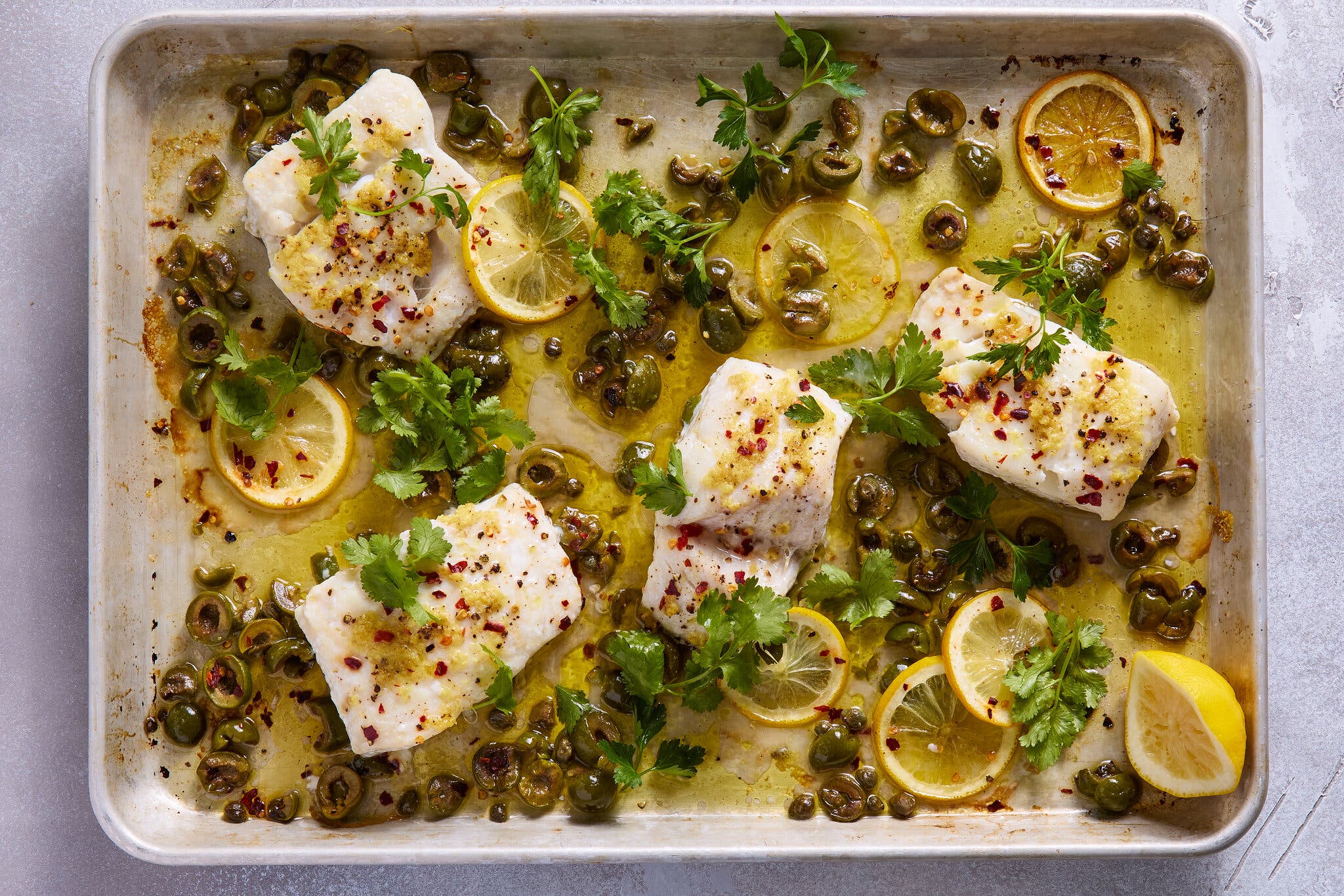 An overhead image of a sheet pan topped with baked fish, olives and lemon wedges.