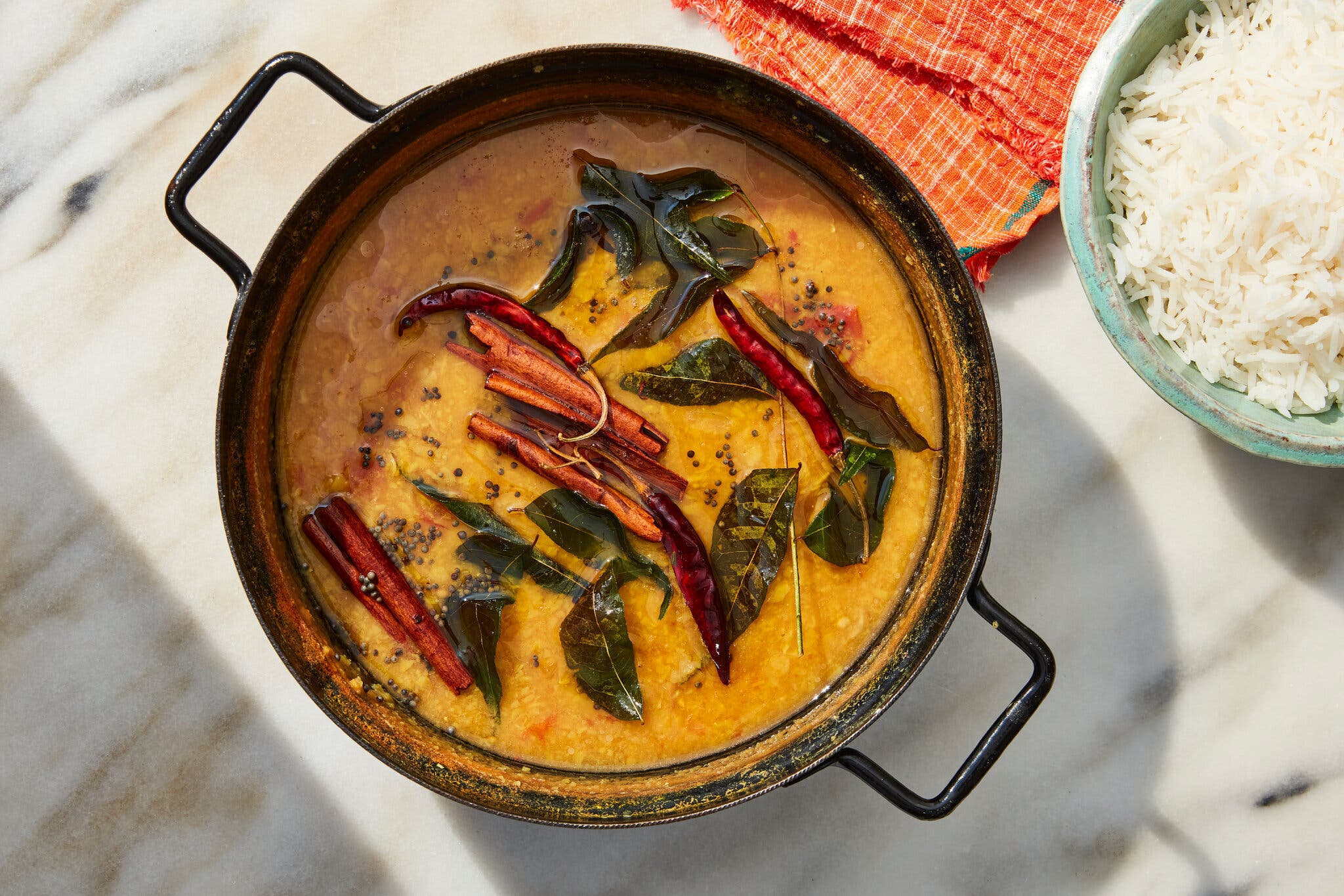 A dark metal pot of yellow lentil dal topped with whole cinnamon sticks, red chilies and curry leaves.
