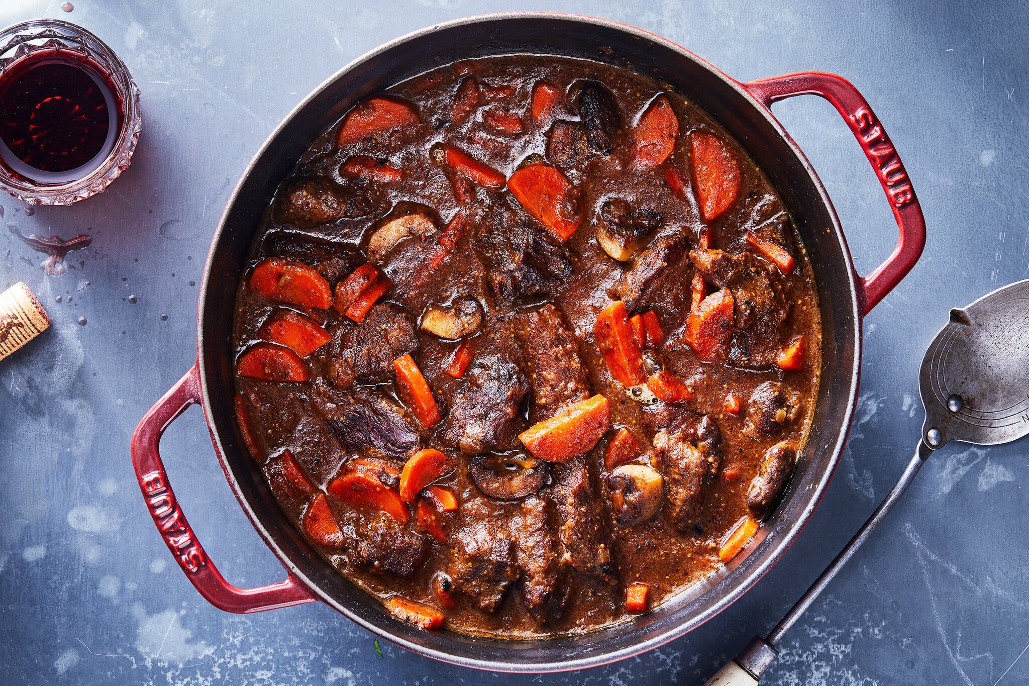 A rich, dark beef stew with sliced carrots and mushrooms in a round red Staub enameled cast-iron pot. A glass of red wine and a spoon sit on the blue tabletop nearby.