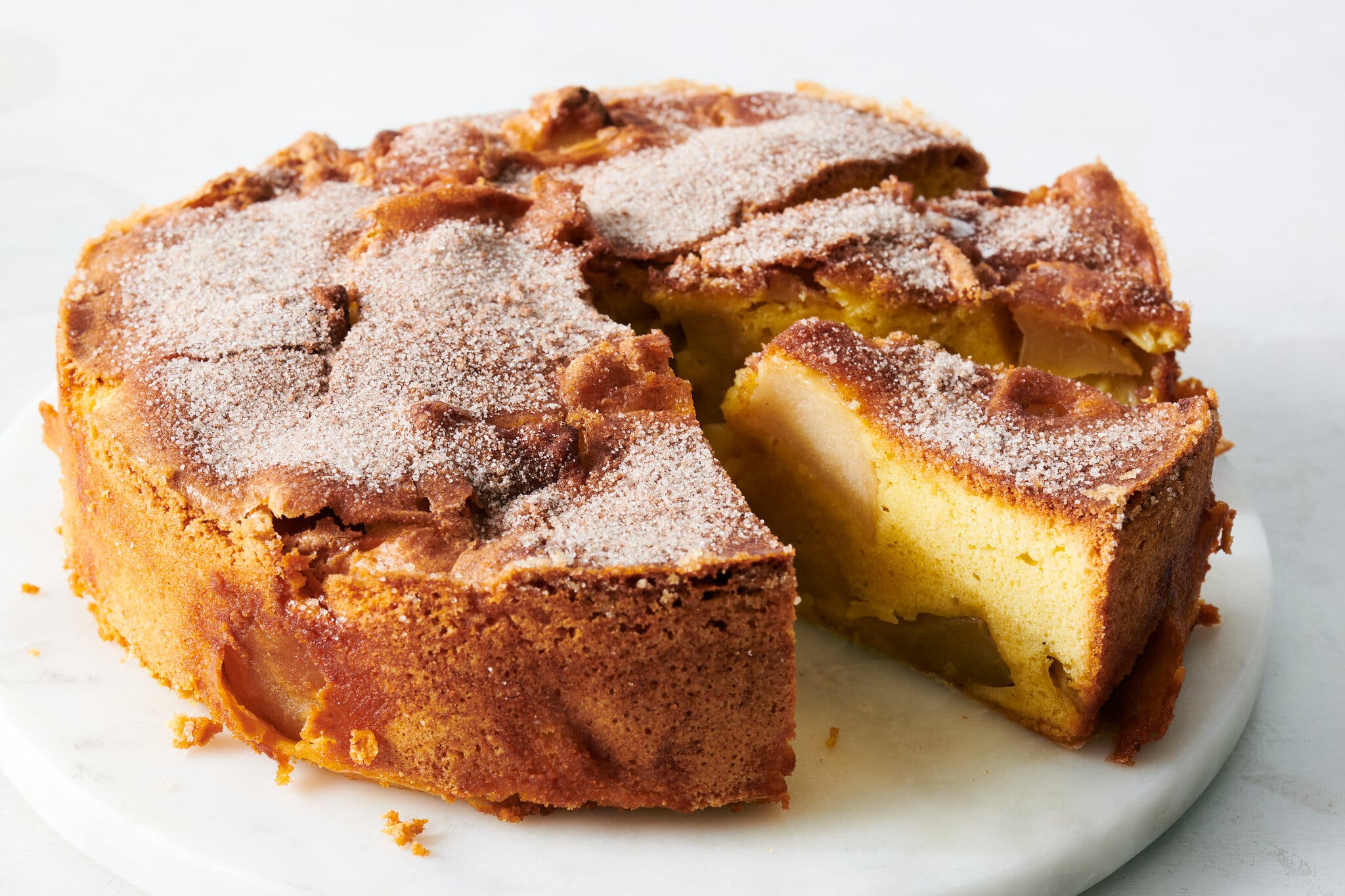 A thick, golden-yellow apple cake with a crackly sugar-dusted top. A large slice is removed, revealing tender chunks of cooked apple embedded in the dense sponge.