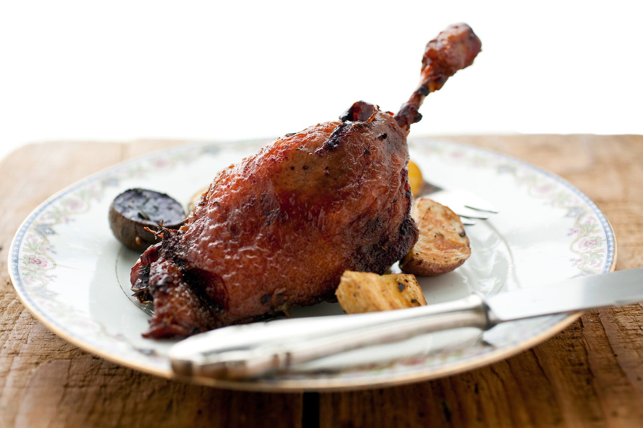 A crisp duck confit leg served with roasted potatoes on a floral rimmed plate.