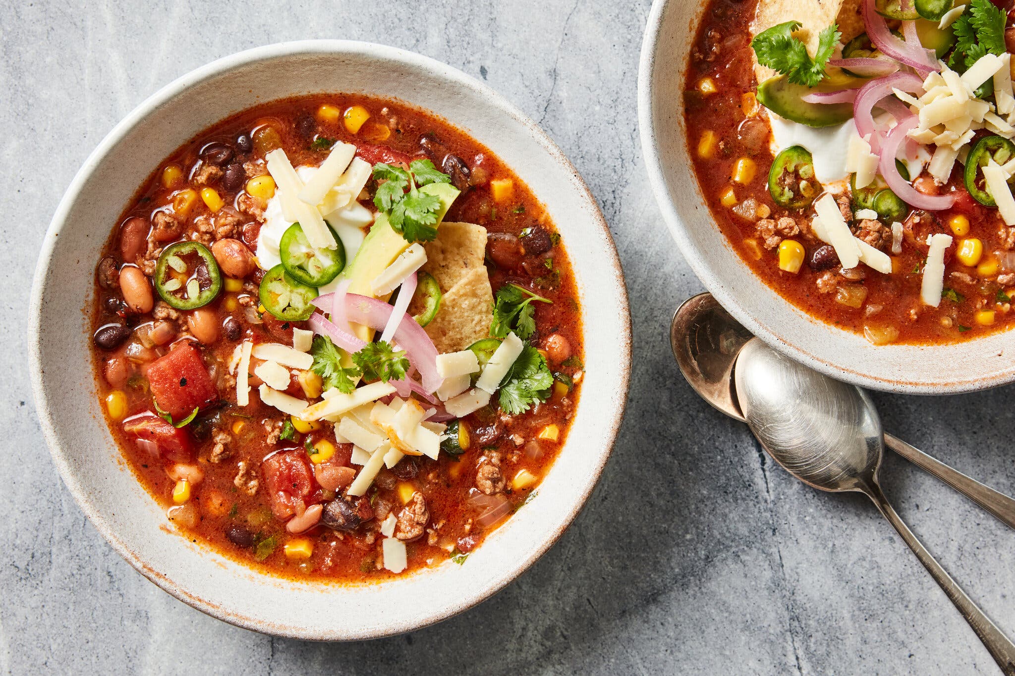 Two white bowls filled with hearty beef and bean soup, topped with avocado, cheese, and red onion.