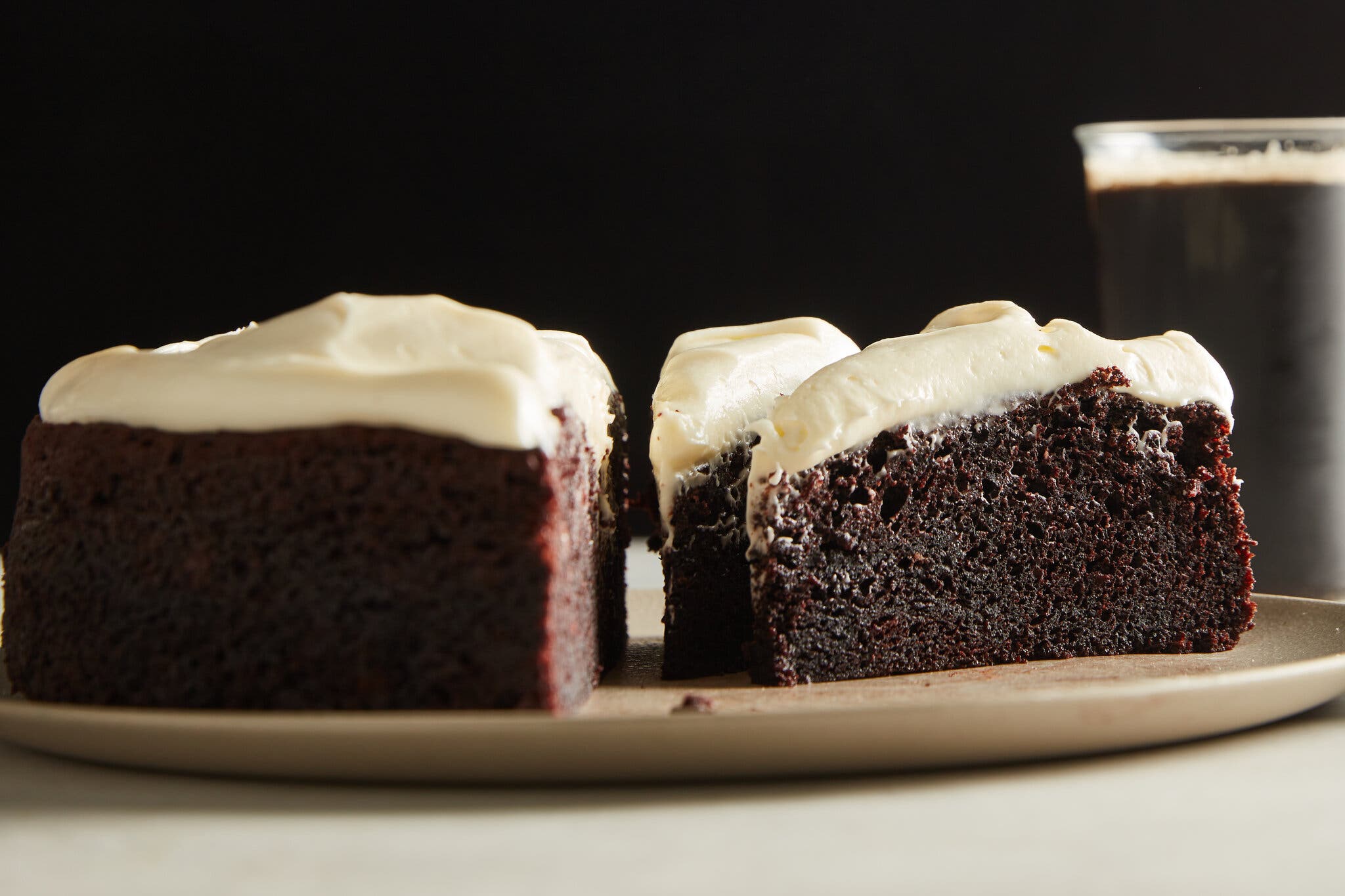 Slices of chocolate Guinness cake with cream-cheese frosting, on a plate. There is a glass of dark stout in the background.