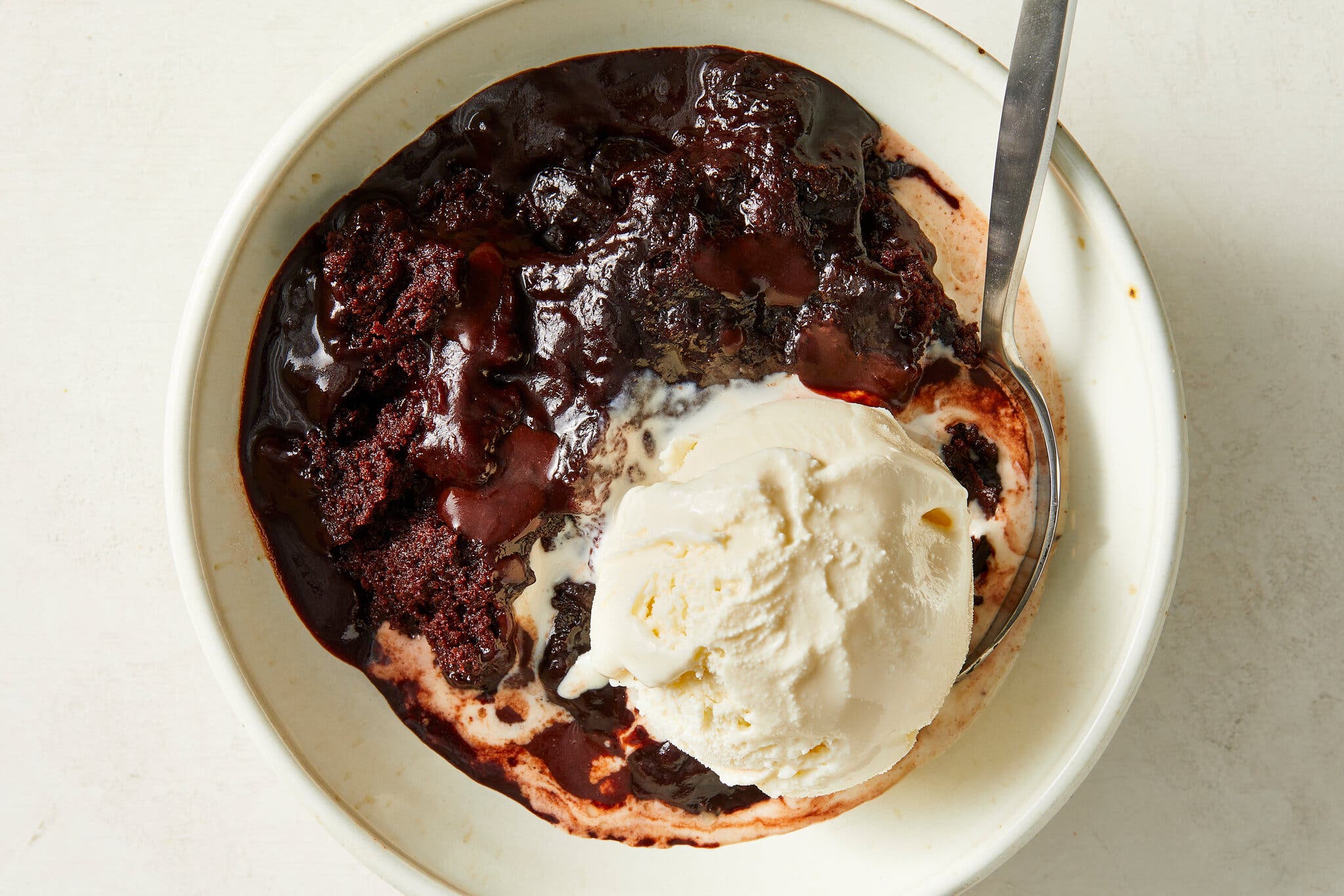 An overhead image of a bowl of chocolate pudding cake, with a scoop of vanilla ice cream on it, slightly melting into the cake. There is a spoon in the bowl.