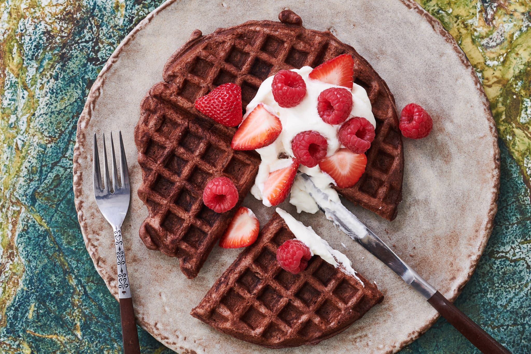 An overhead image of a chocolate waffle topped with whipped cream, strawberries and raspberries, cut into triangular pieces on a serving plate. There are a fork and knife cutting into the waffle.