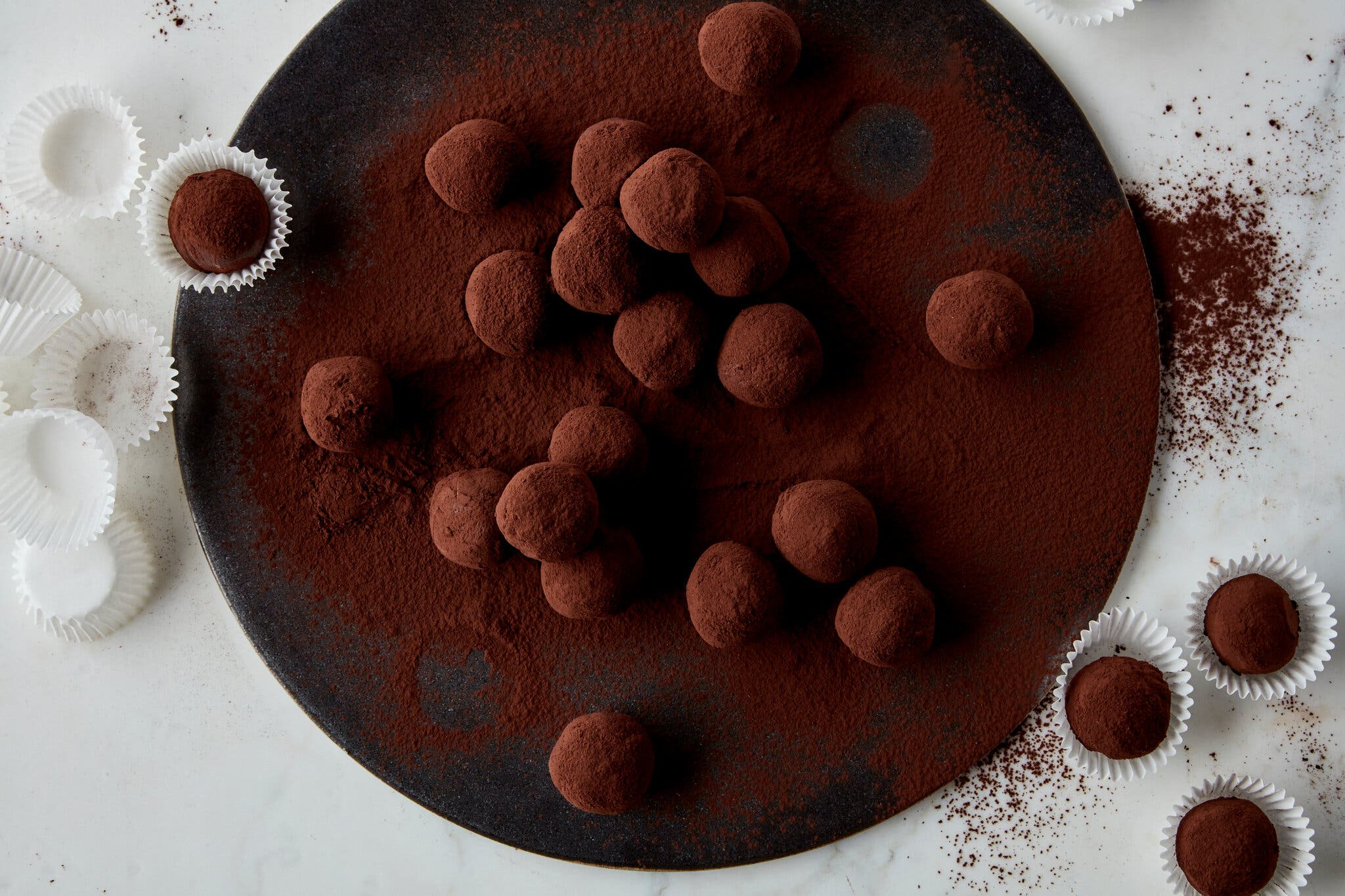 An overhead image of a black plate of chocolate truffles, dusted in cocoa powder. Some are in paper wrappers.
