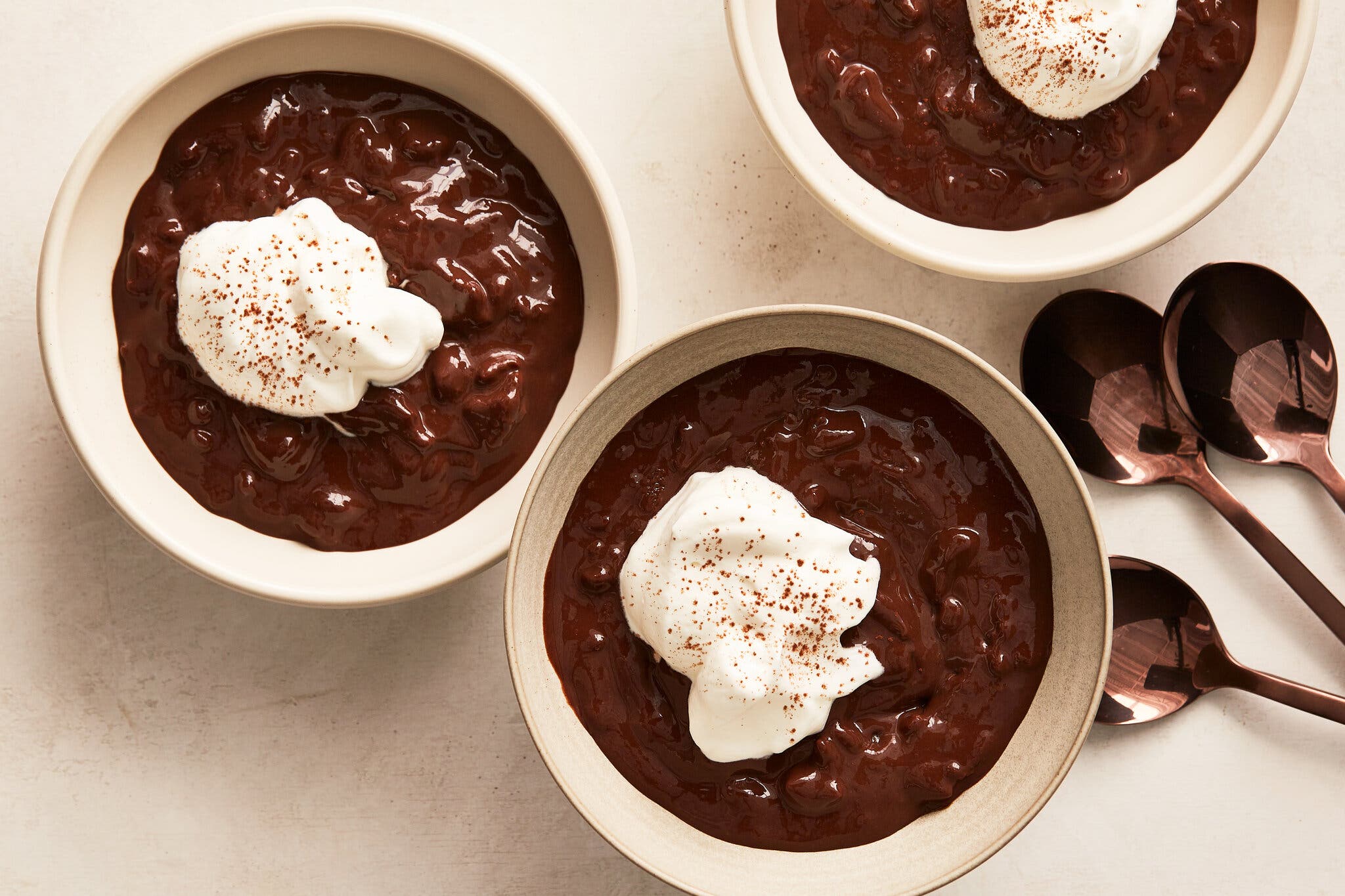 An overhead image of three bowls of glossy chocolate rice pudding, topped with whipped cream and shaved chocolate. There are three spoons nearby.