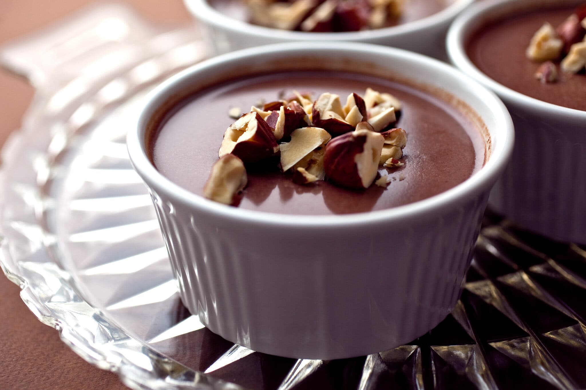 A ramekin of bouncy chocolate panna cotta topped with chopped hazelnuts, on a glass serving plate.